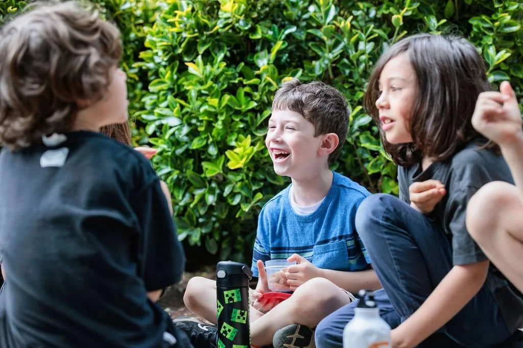 Four children sitting outdoors and laughing with each other, surrounded by greenery.