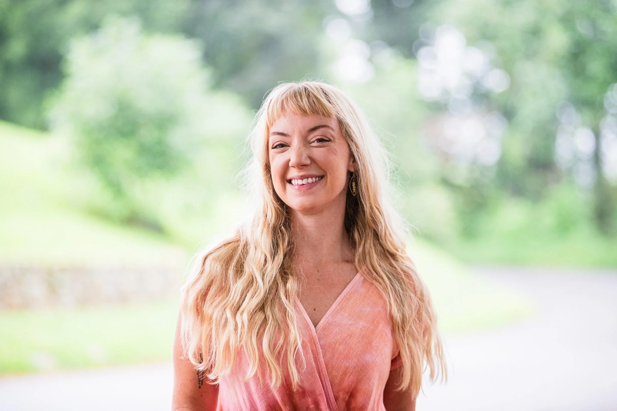 A young woman with long, wavy blonde hair wearing a pink top, smiling, standing outdoors with a blurred green background.