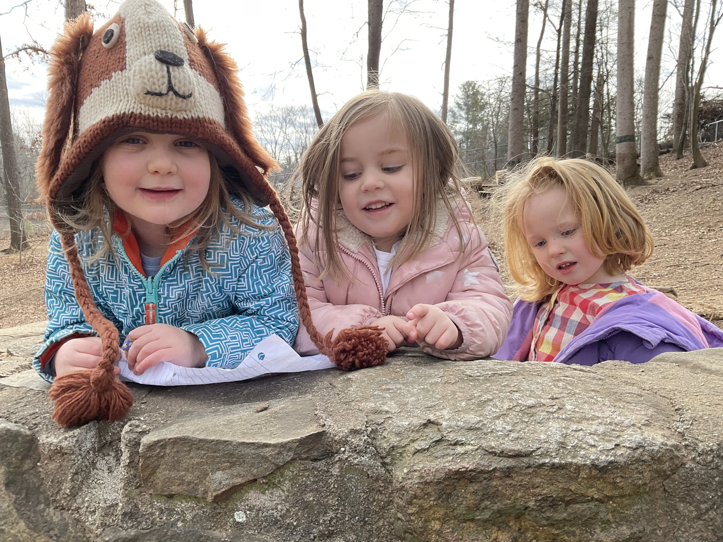 Three young girls leaning on a large rock outdoors in a wooded area, one wearing a lion hat, and the other two in pink and purple jackets.
