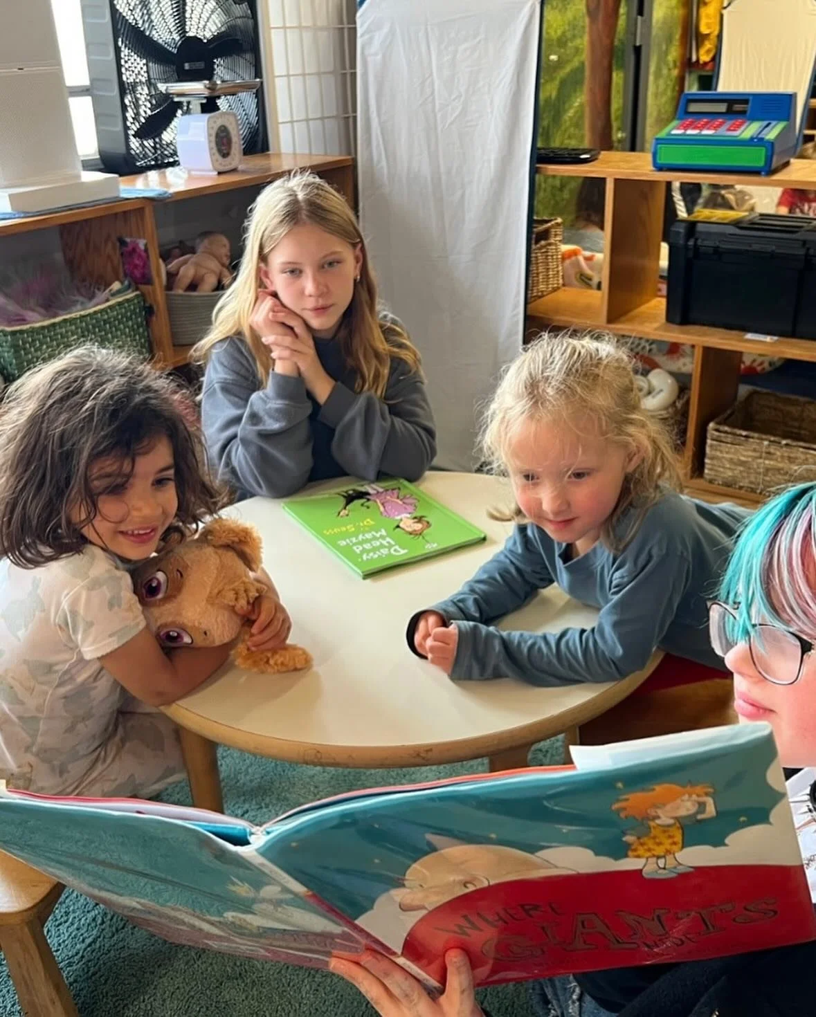 a middle schooler with colored hair reading a picture book to a group of curious preschoolers