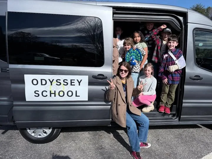 A grey van with a sign for Odyssey School on it with lots of kids smiling from the open door