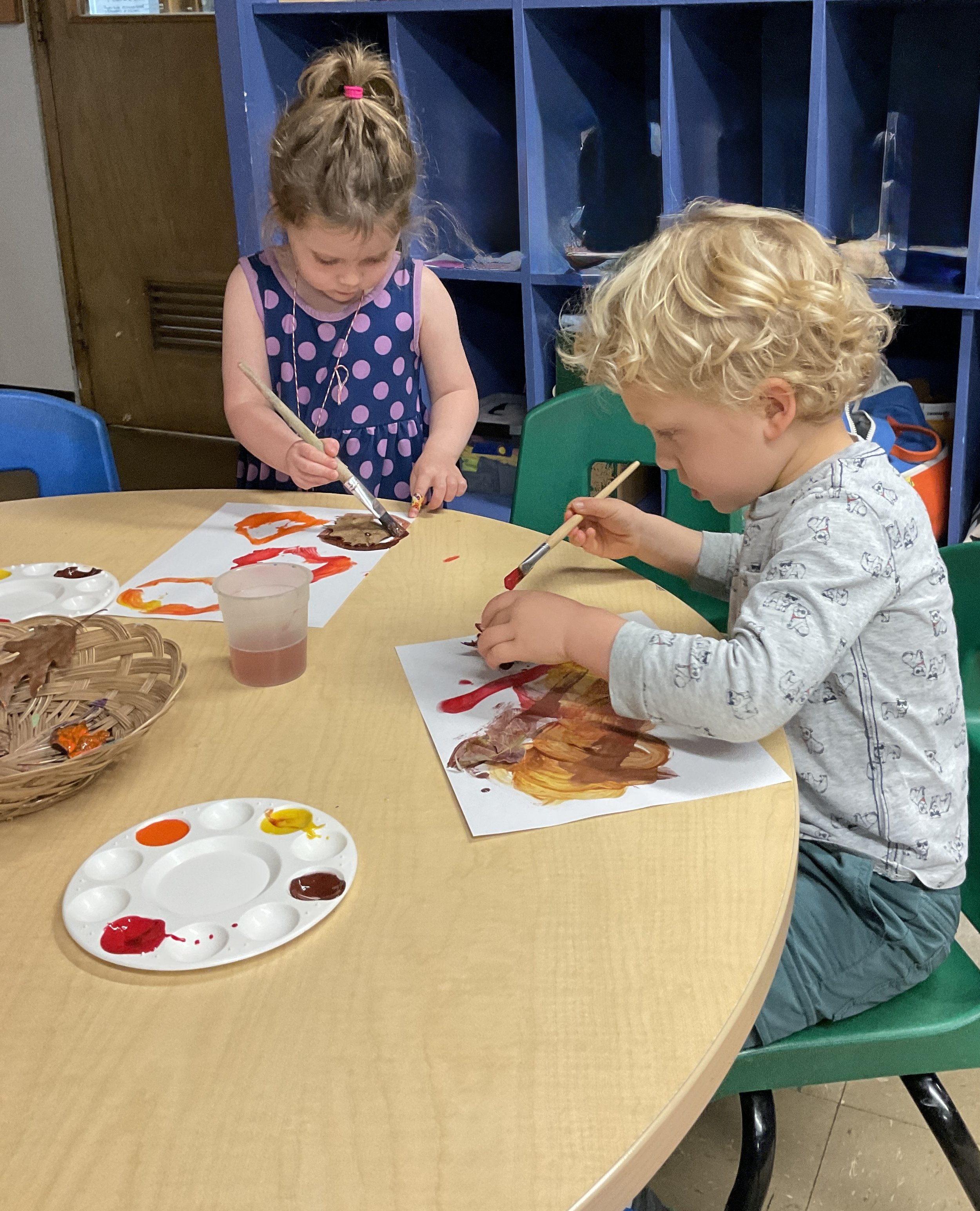 Two young children painting with watercolors at a round table. The girl is standing and wearing a purple polka dot dress, and the boy is sitting in a green chair, wearing a gray shirt with small cartoon characters. They are focused on their artwork, 