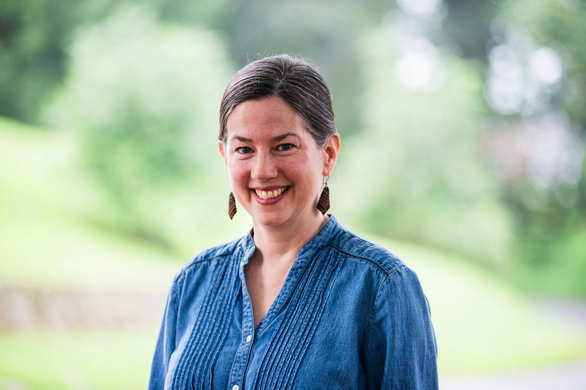 A woman with dark hair pulled back, wearing earrings and a blue button-up shirt, smiling outdoors with a blurred green background.