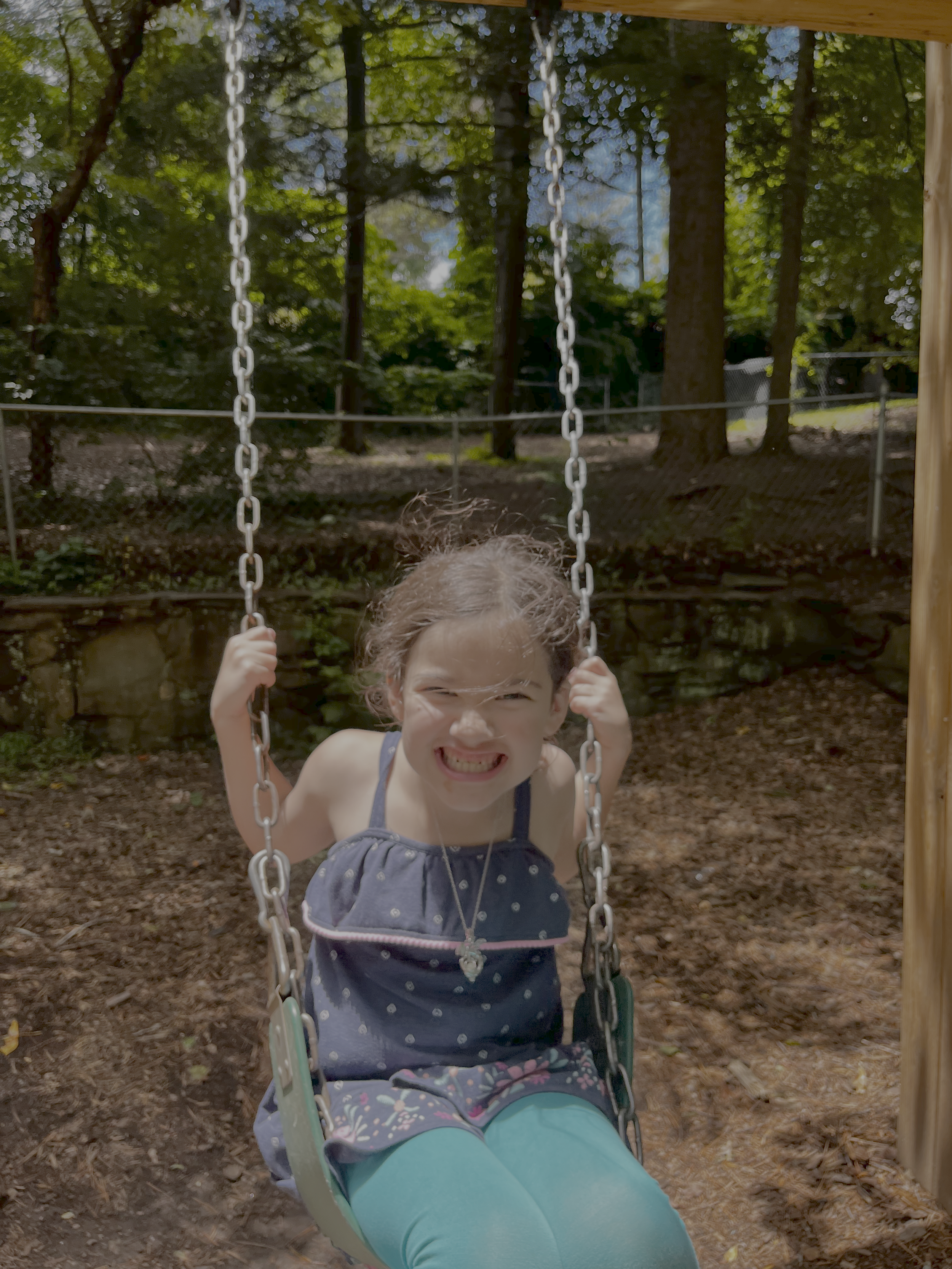 Young girl on a swing smiling and making a playful face outdoors in a wooded playground at summer camp.