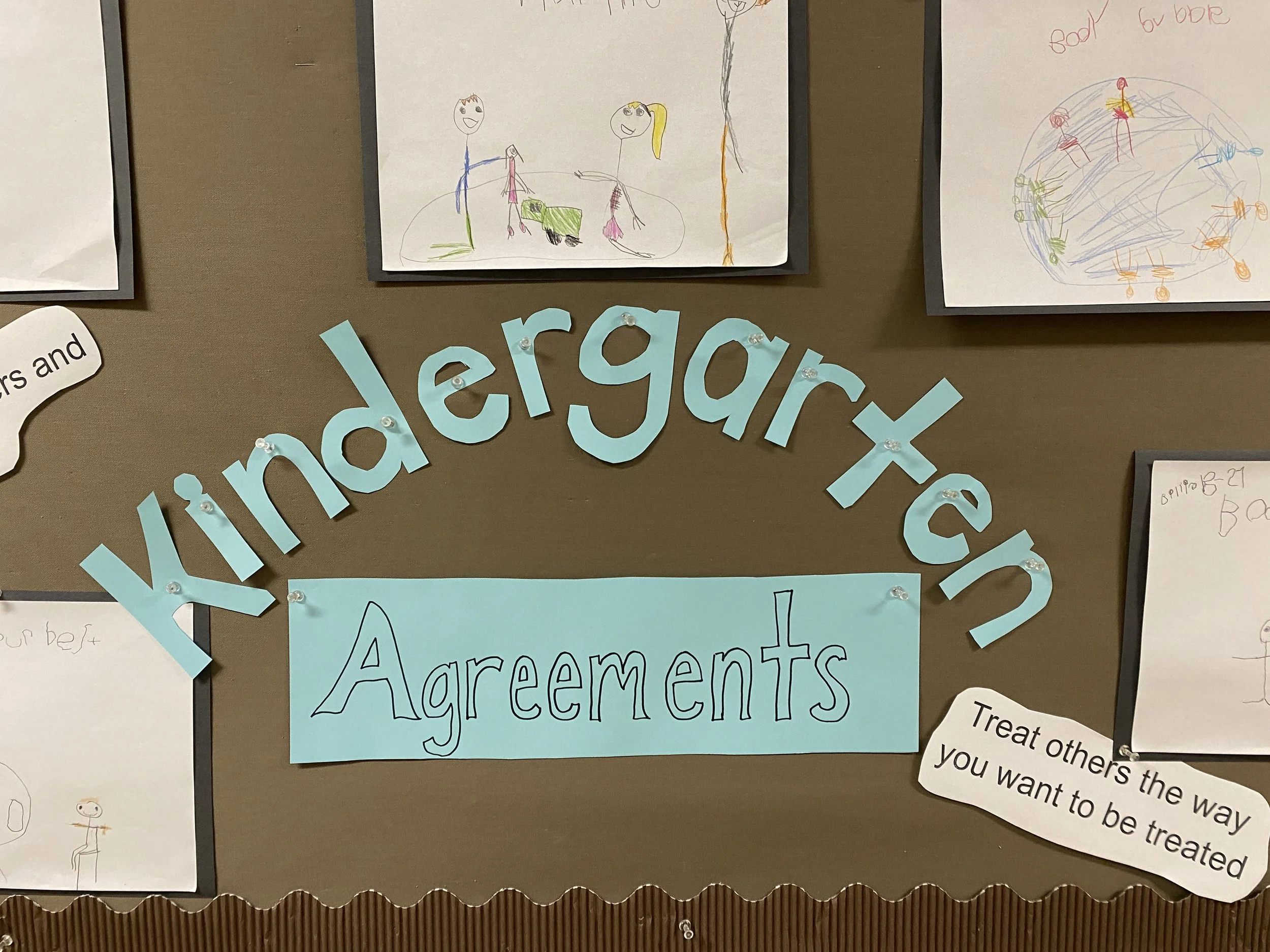 A bulletin board with the words 'kindergarten agreements' in large blue cutout letters and a blue paper sign with the word 'Agreements' written on it. Surrounding the text are children's drawings on white paper, some with colorful figures and objects, and a white paper with a quote about treating others well.