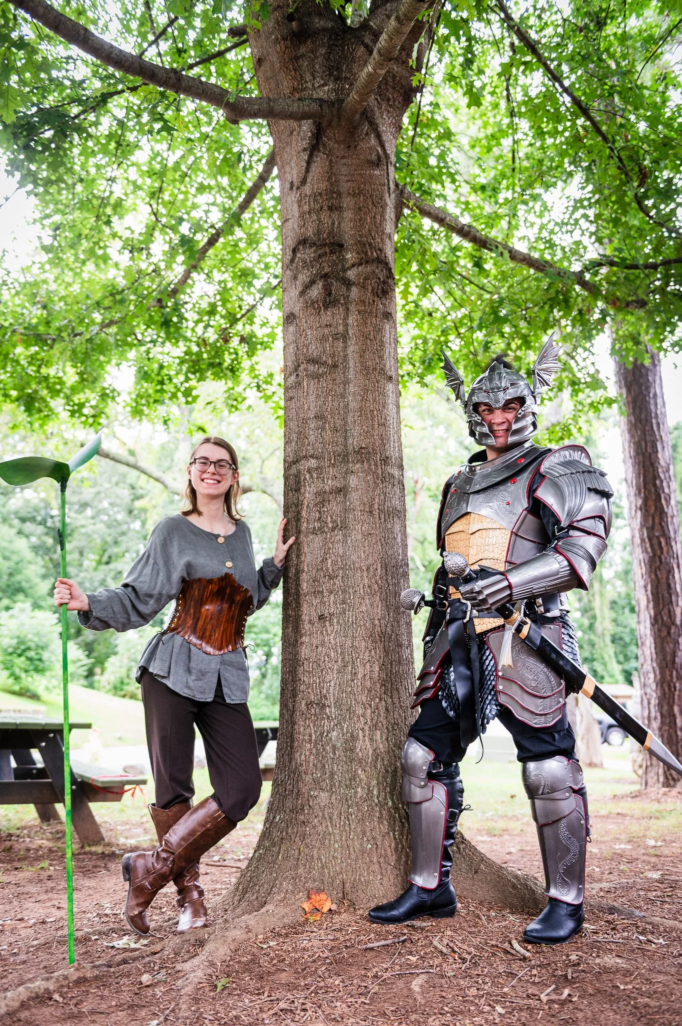 A woman dressed in casual medieval-inspired clothing and a man in detailed knight armor stand outdoors in a park, posing next to a large tree with lush green leaves. The woman holds a green staff with a decorative top, smiling at the camera, while th