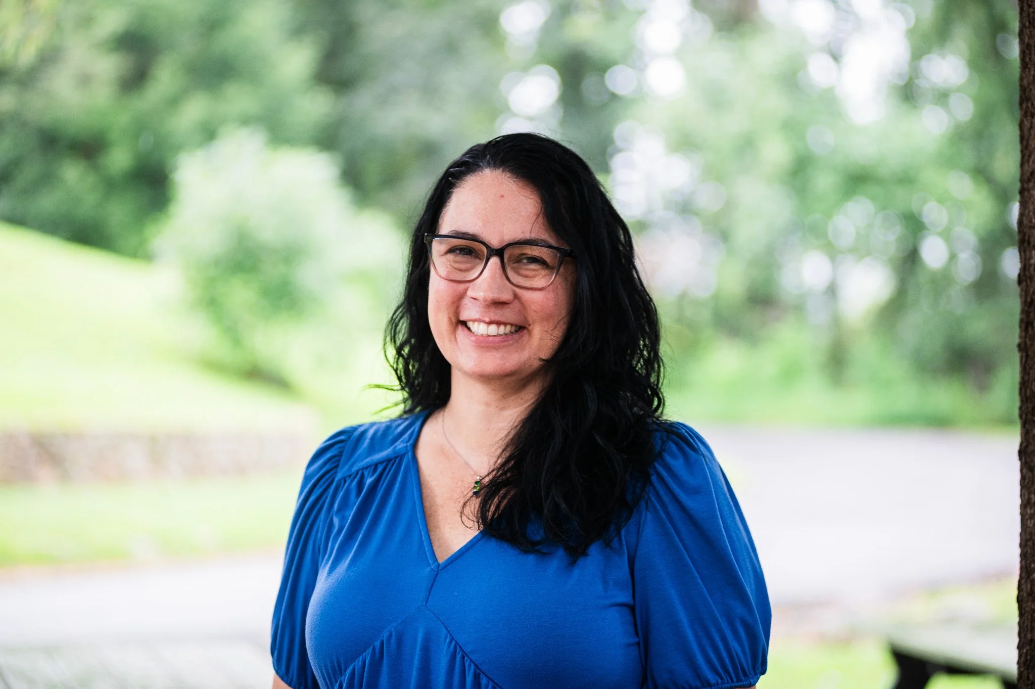 a portrait of a woman with shoulder length dark hair and glasses and a blue shirt posing against a blurred forest background