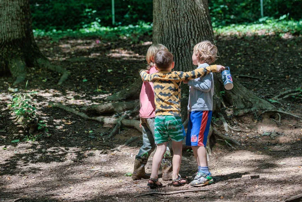 Three children hugging in a wooded forest playground, standing by a tree, holding a water bottle.