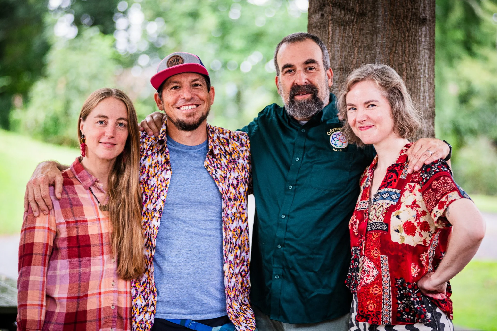 Four people stand outdoors beneath a tree, with three men and one woman. They are smiling and have their arms around each other, wearing casual, colorful clothing.
