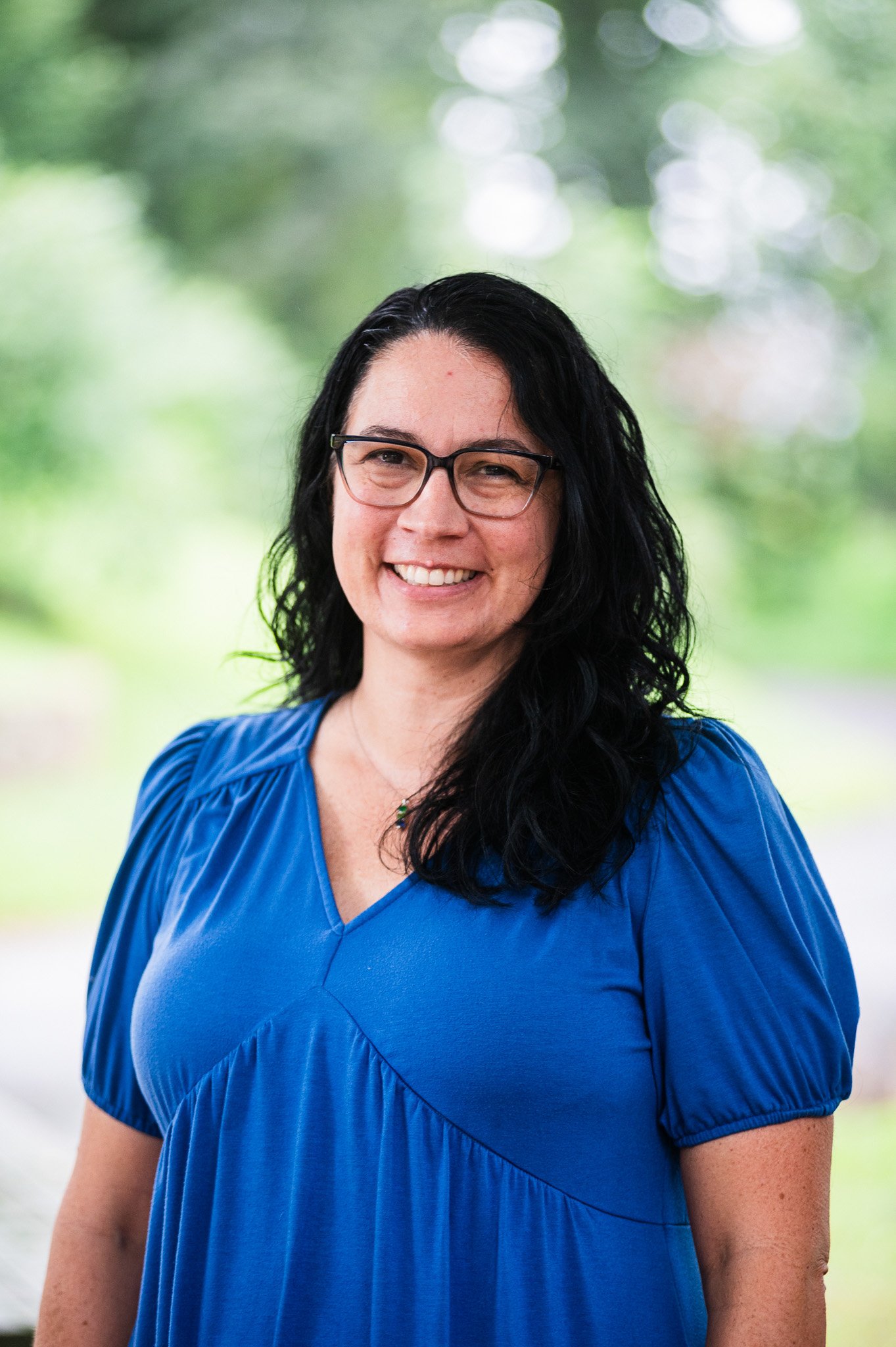 A woman with wavy black hair, a blue top, and glasses smiling for the camera on a forested background
