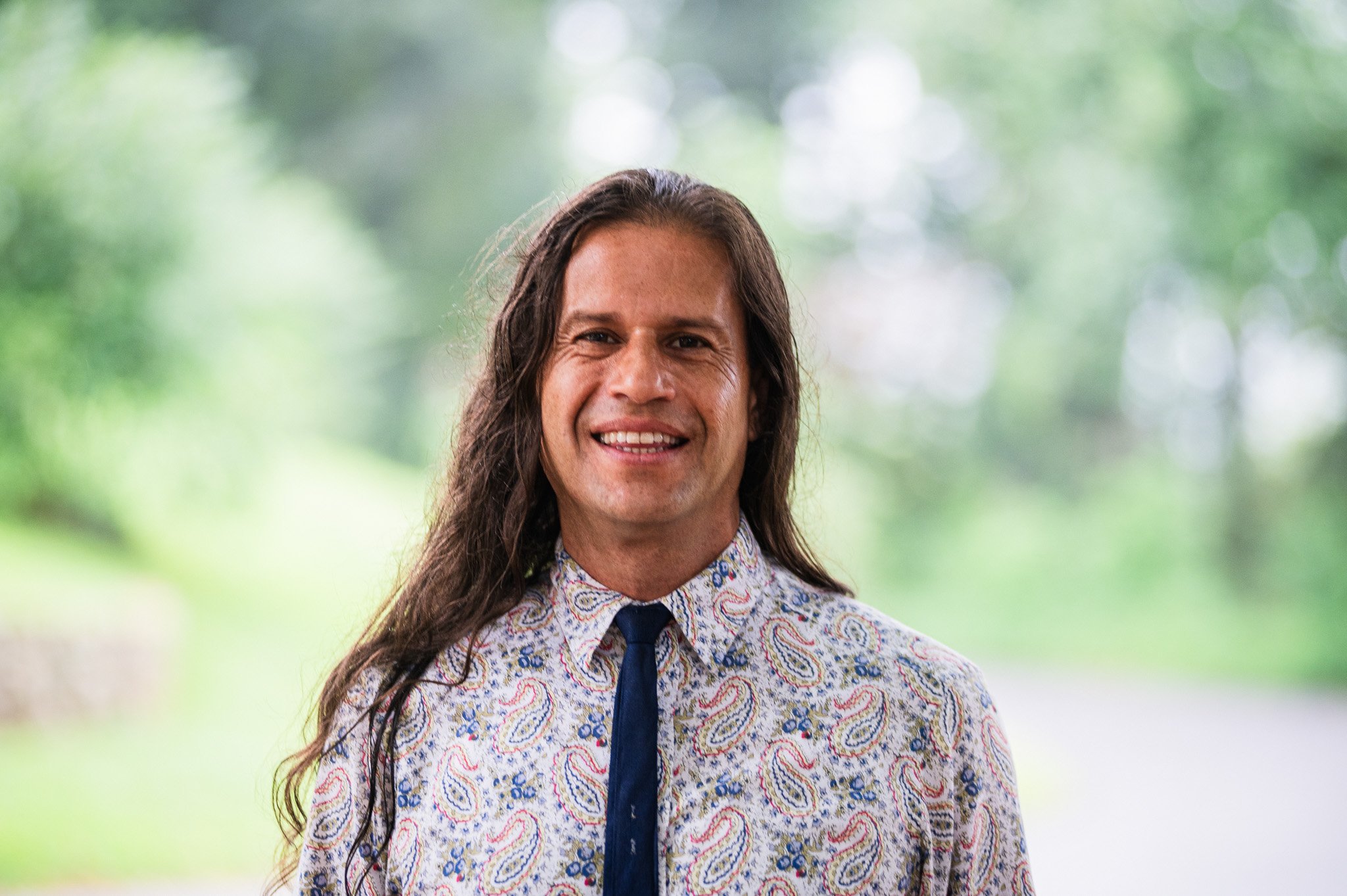 A man with long brown hair smiling outdoors, wearing a light-colored shirt with a paisley pattern and a dark tie, with a blurred green natural background.