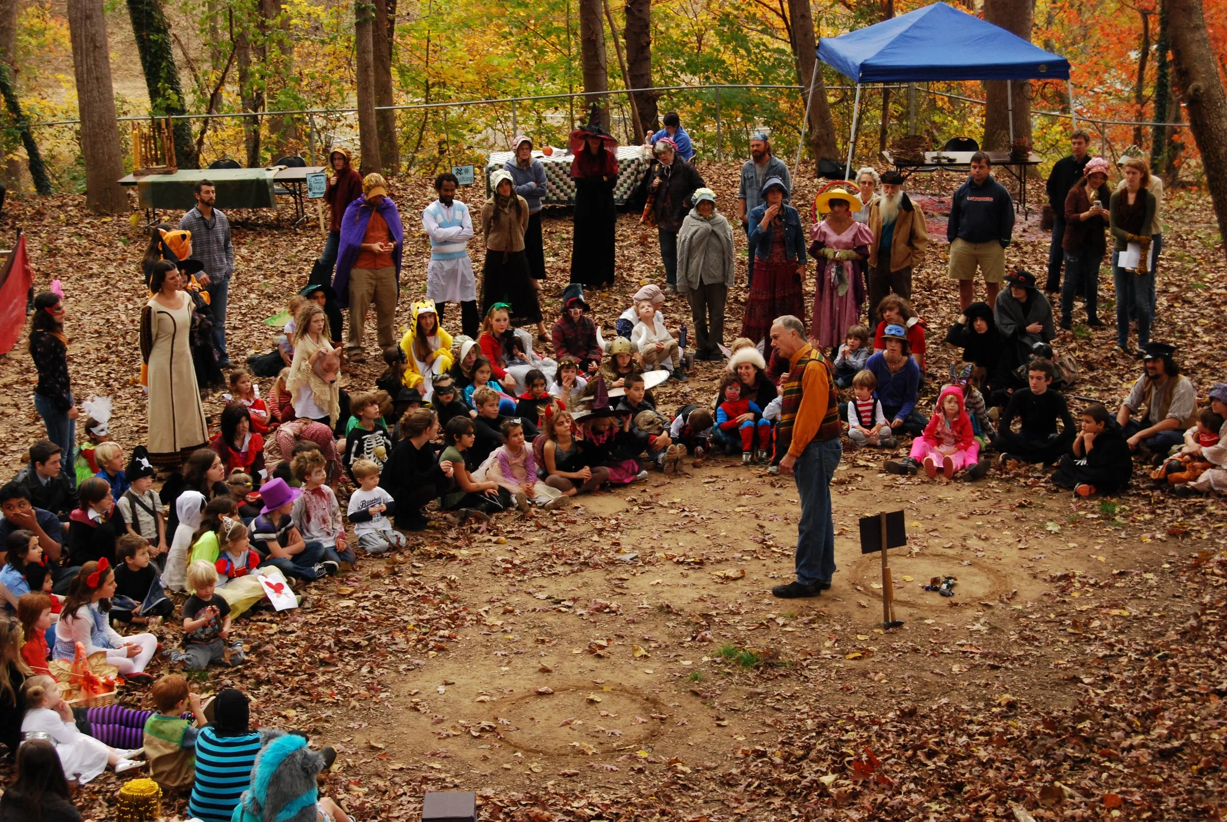 Group of children and adults gathered outdoors in a wooded area during autumn, with many dressed in costumes, sitting on leaves listening to a man speak in front of them.