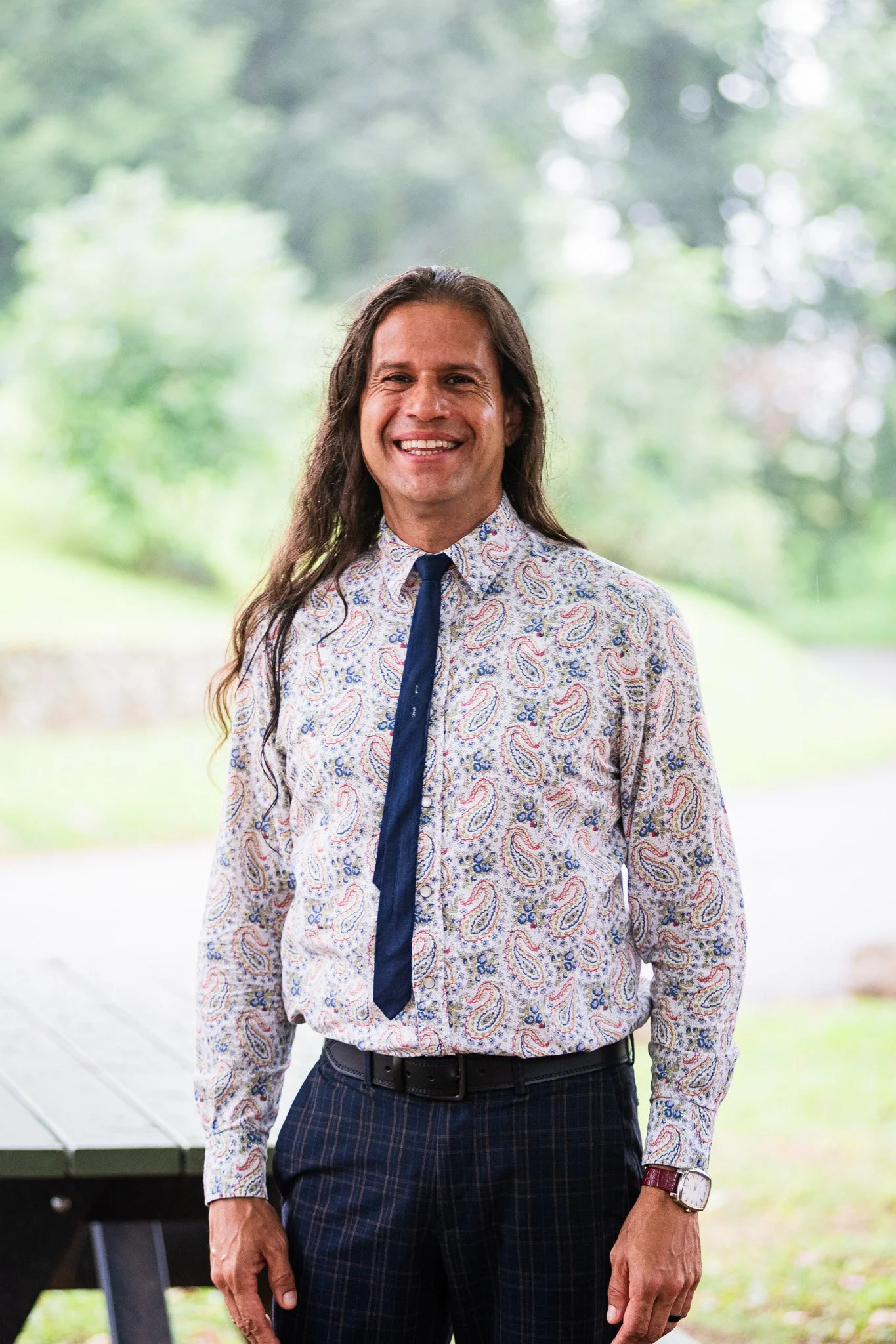 A man with long dark hair, wearing a colorful paisley shirt, dark plaid pants, and a dark tie, smiling outdoors with a blurred natural background.