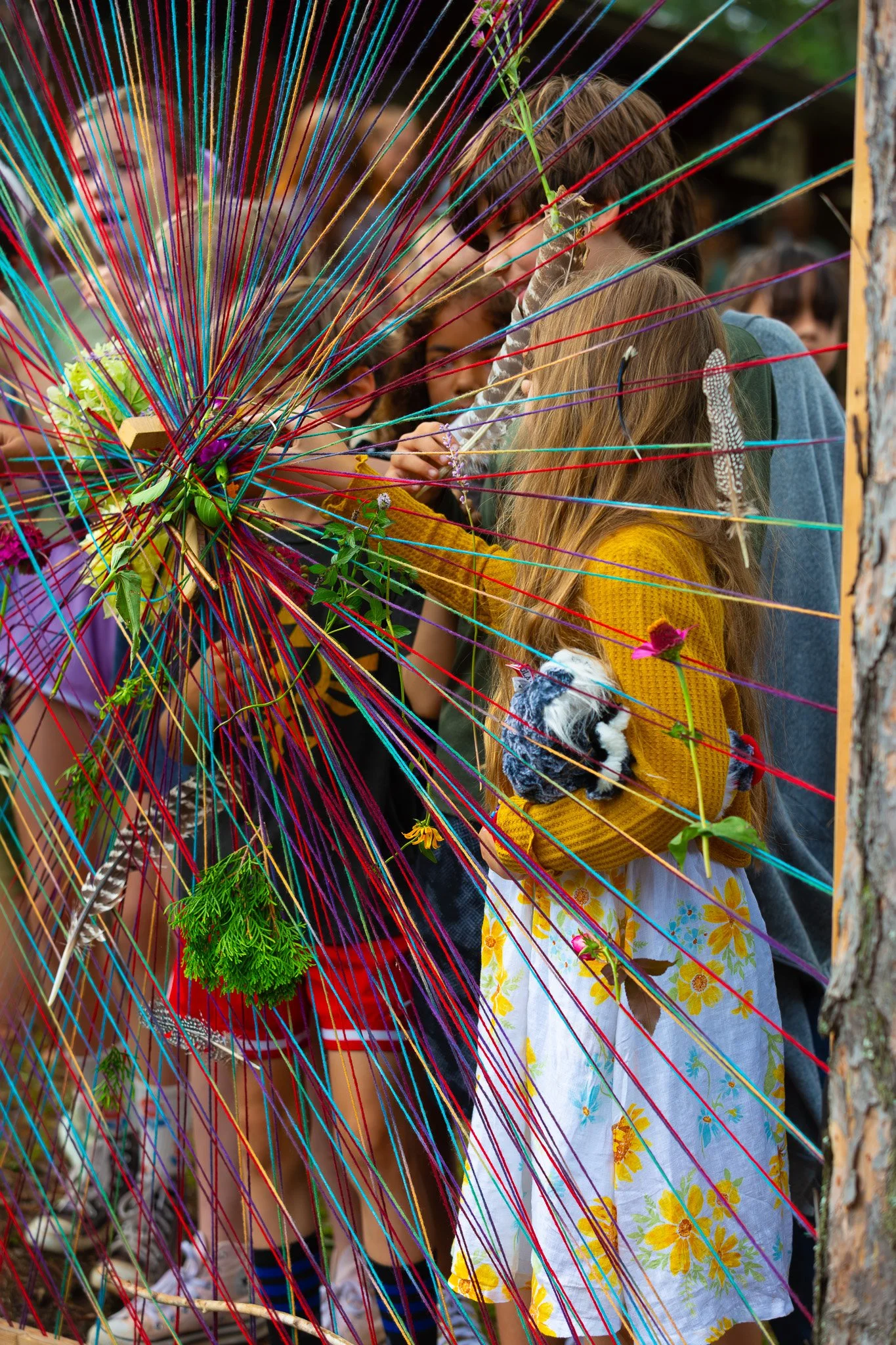 Children gather behind a large, colorful, spoked art installation made of numerous threads in various colors, with some children touching or looking through it.