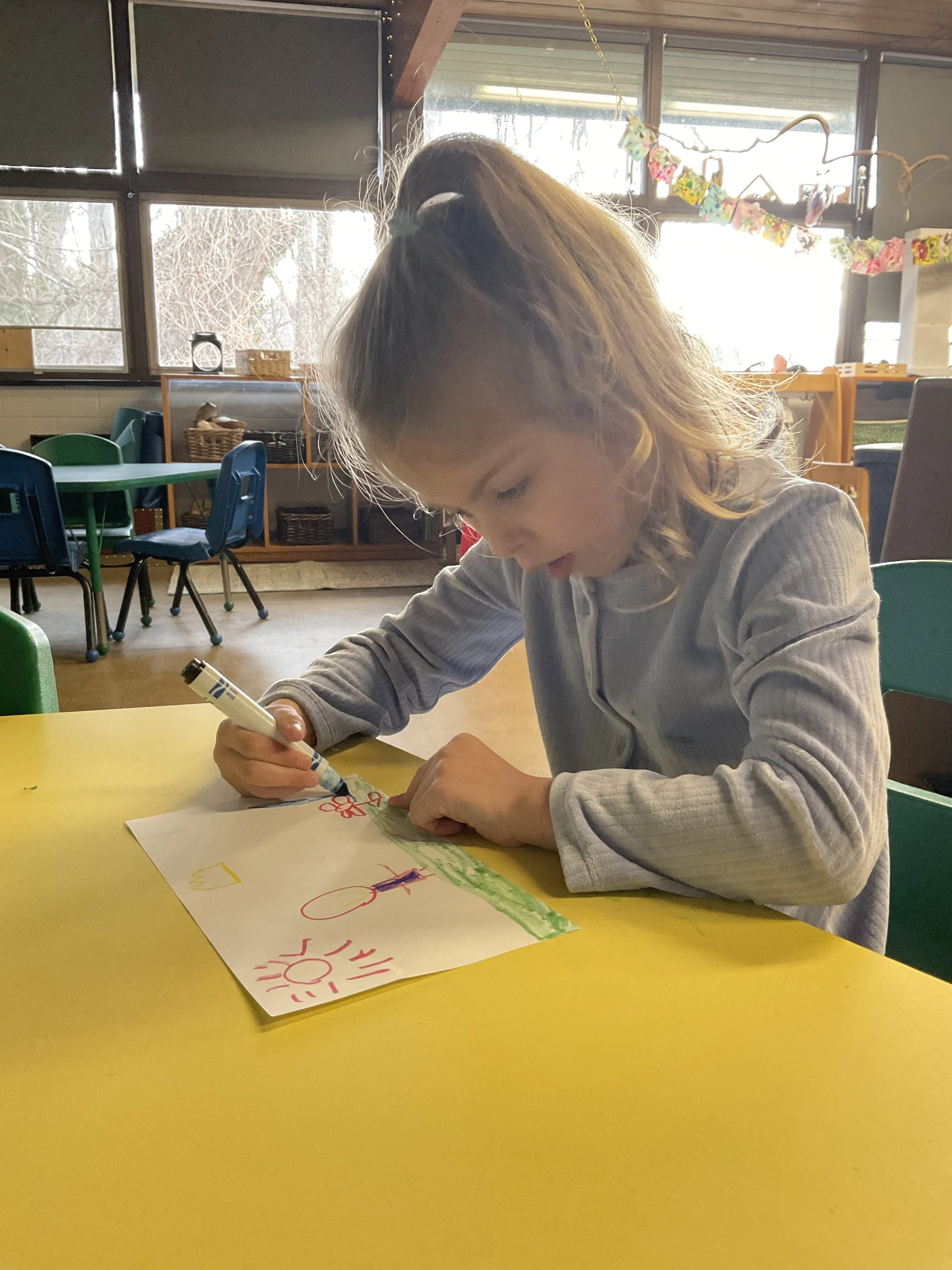A young girl with brown hair tied in a high ponytail, drawing with markers on a white sheet of paper at a yellow table in a classroom.