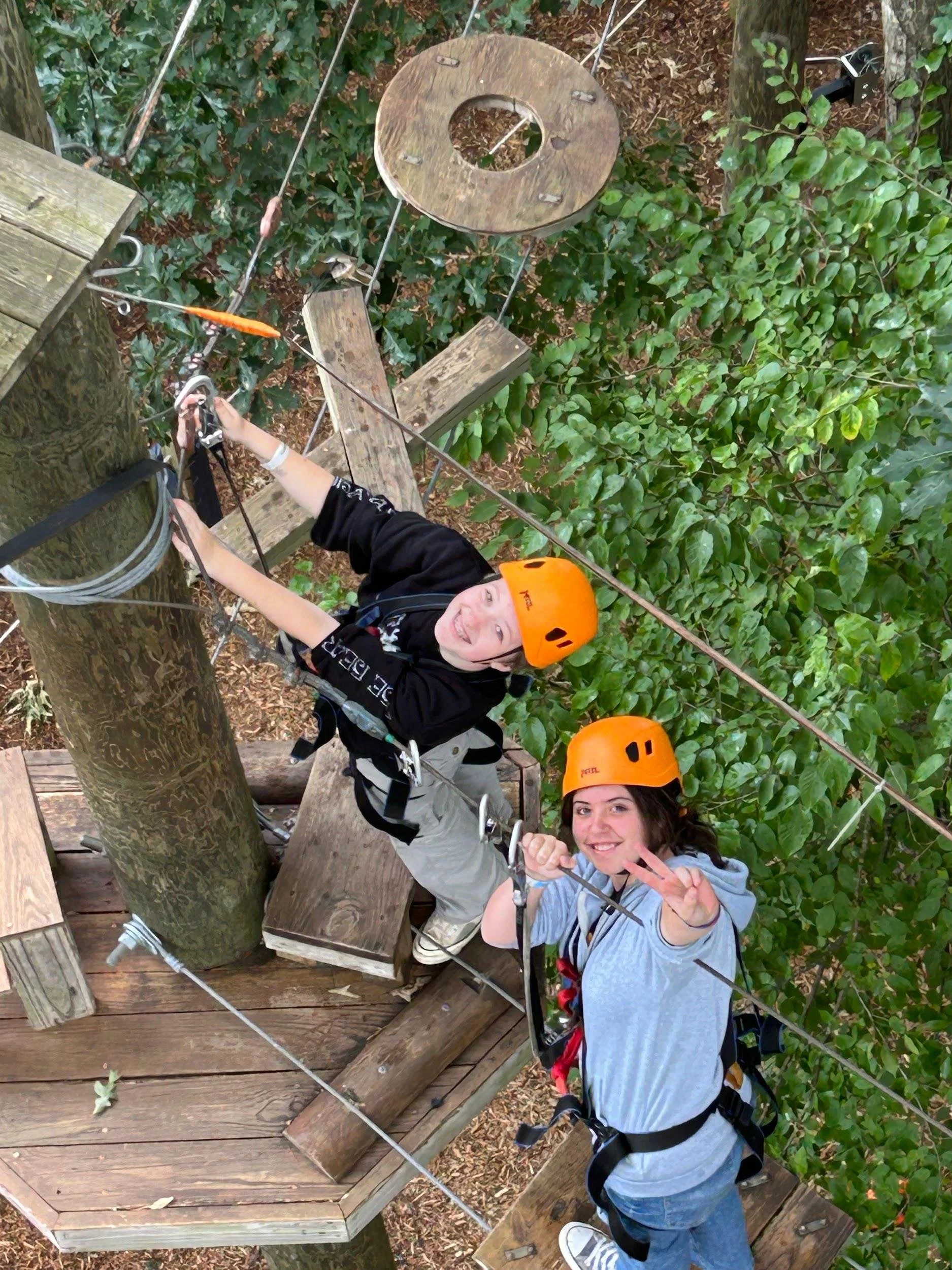 two high school girls in orange helmets smiling at the camera above them on a ropes course