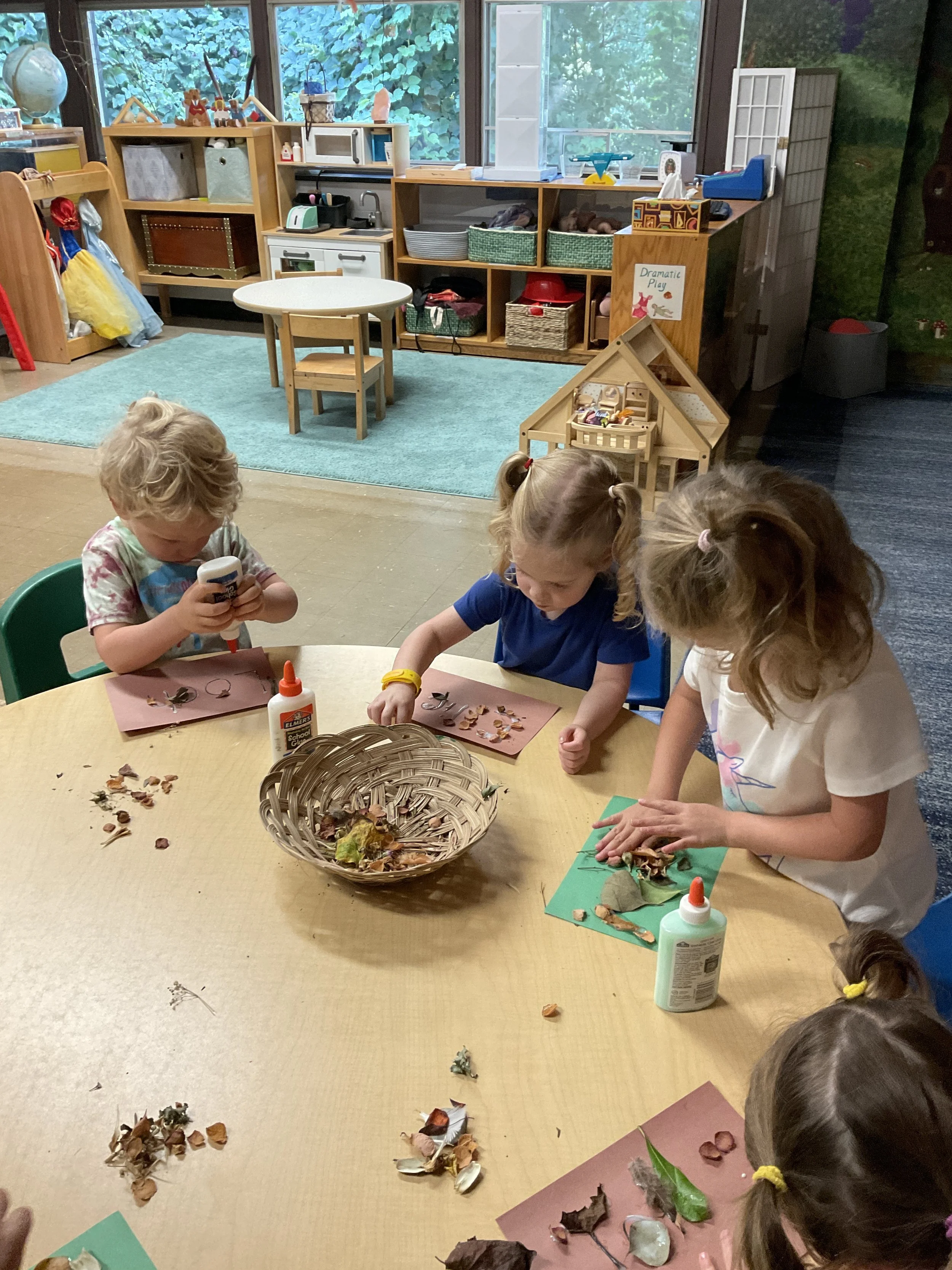 Three children engaged in a craft activity at a round table in a classroom, using glue and natural objects like leaves and shells on colored mats.