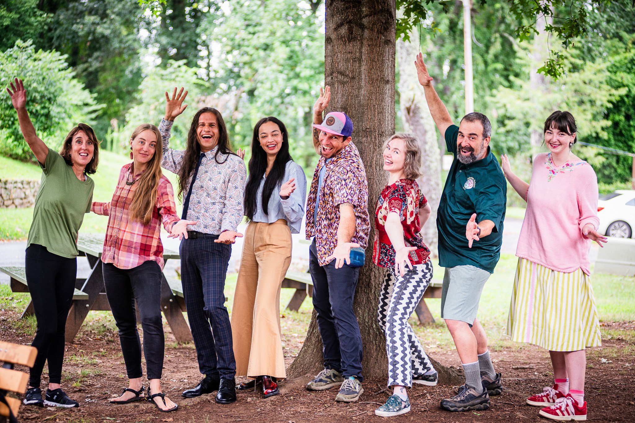 A group of nine diverse people standing outdoors under a tree, smiling, and posing with outstretched arms for a group photo.