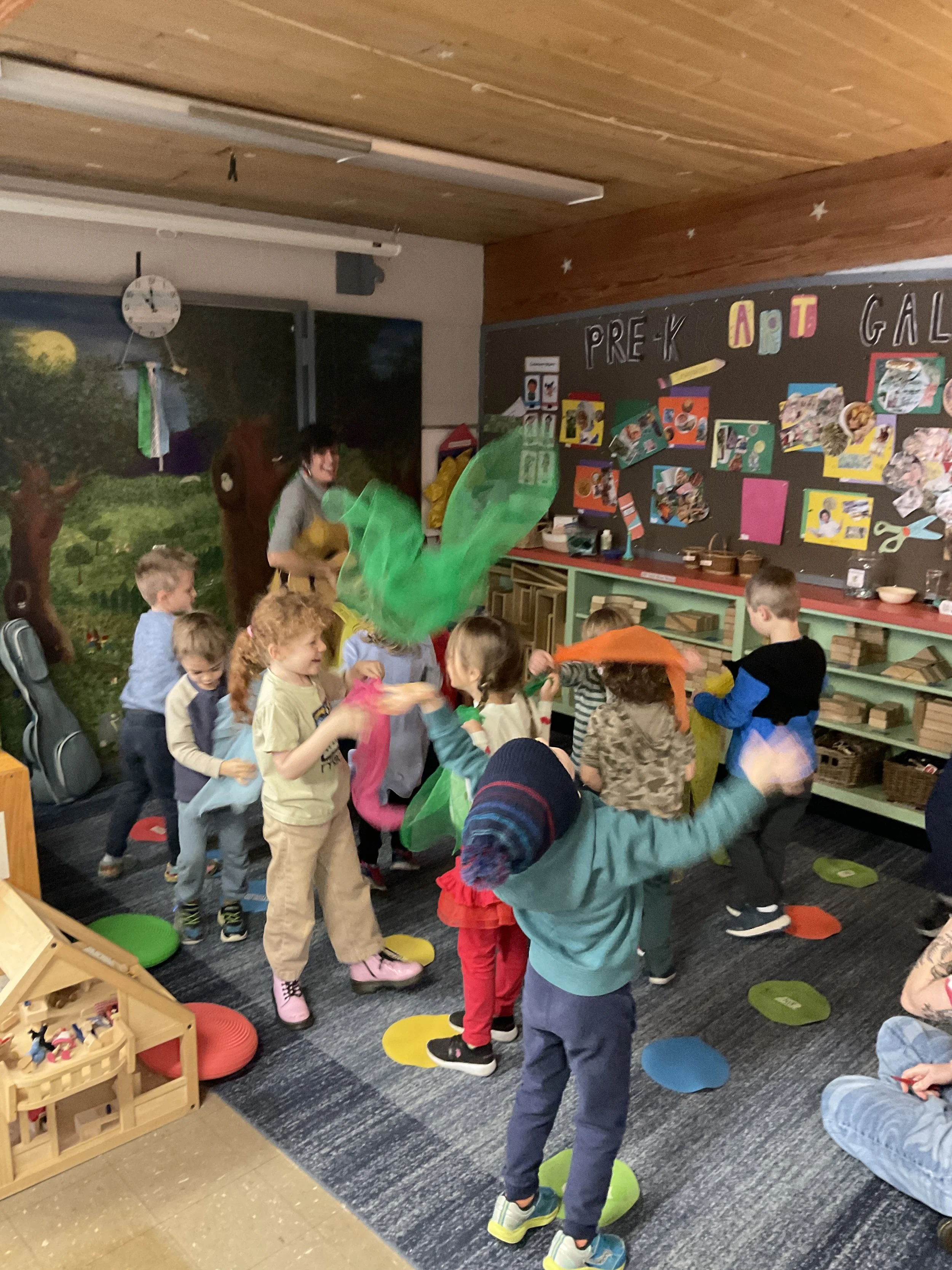 Children participating in a Spanish class game involving colorful cloth ribbons in a decorated classroom with the sign 'Pre-K Art Gallery' on the wall.