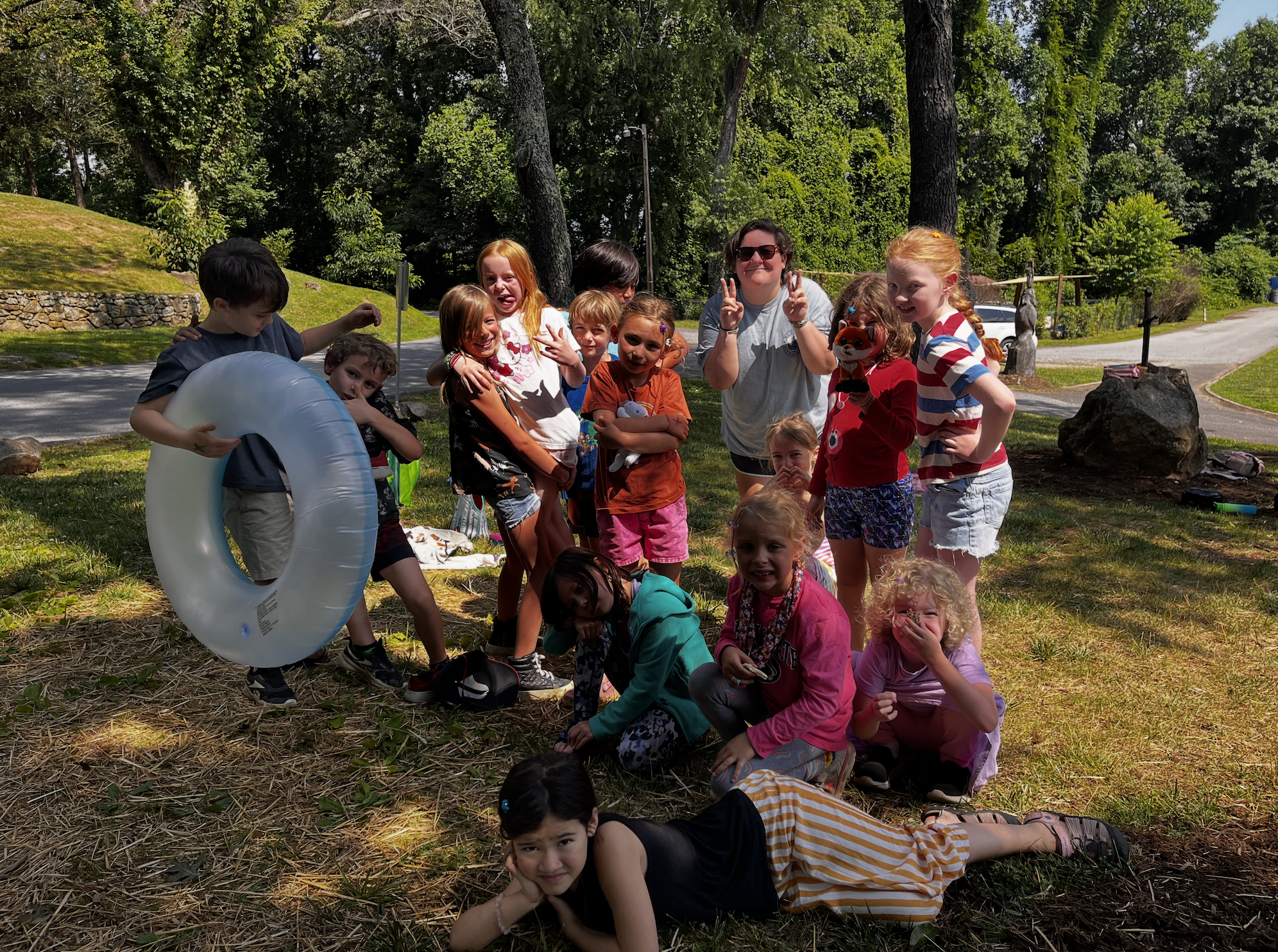 A group of children and two adults pose for a photo outdoors, surrounded by trees and grass. Some children are standing, others are sitting or lying on the ground, and one is holding a floatation ring. The children are smiling and making playful gestures.