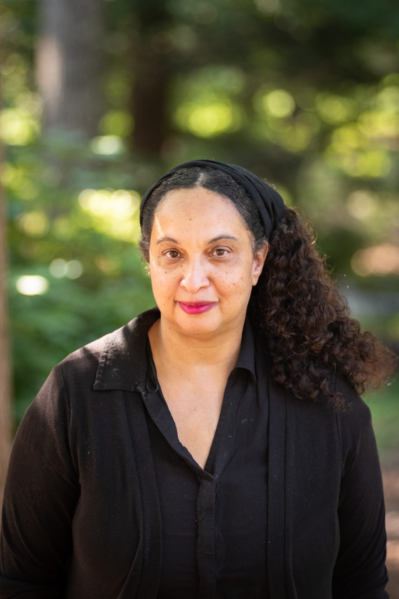 A woman with curly dark hair wearing a black headband and black shirt stands outdoors with a blurred green forest background.