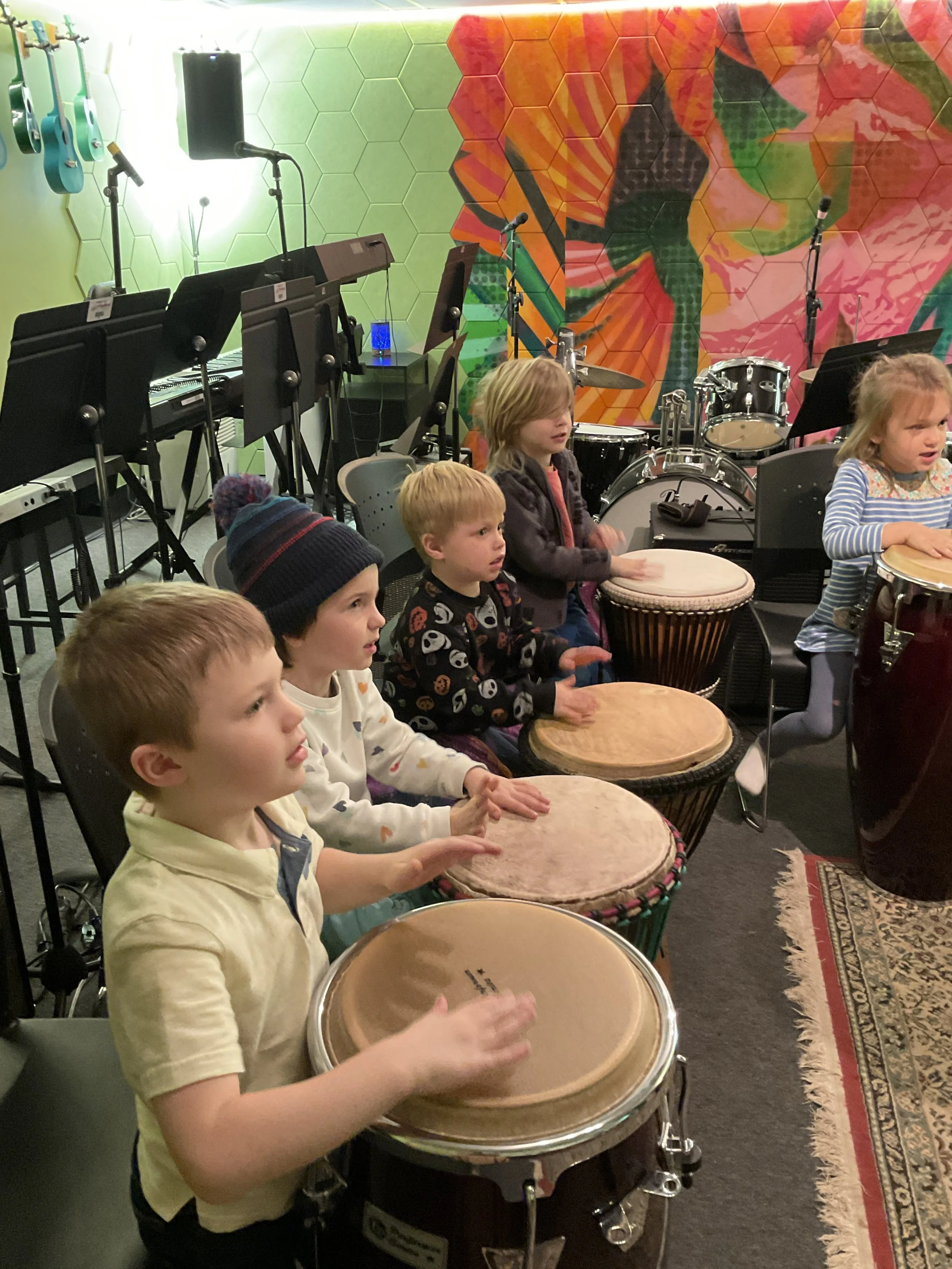 Group of young children sitting and playing drums in a music room, with musical instruments and a colorful abstract wall mural in the background.
