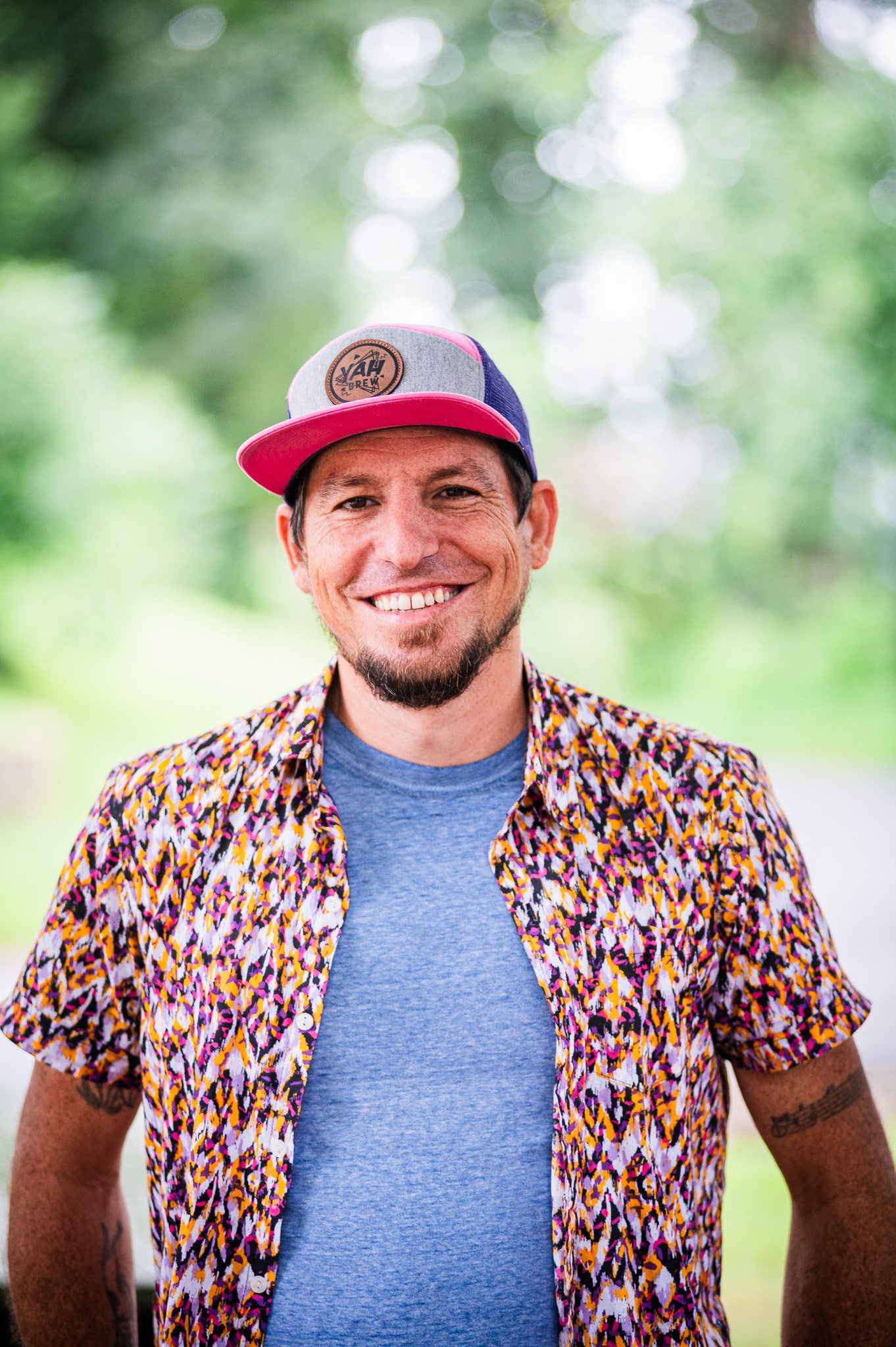 a man with a goatee, a flat brim cap, and a colorful button down open over a blue tee smiling on a wooded background