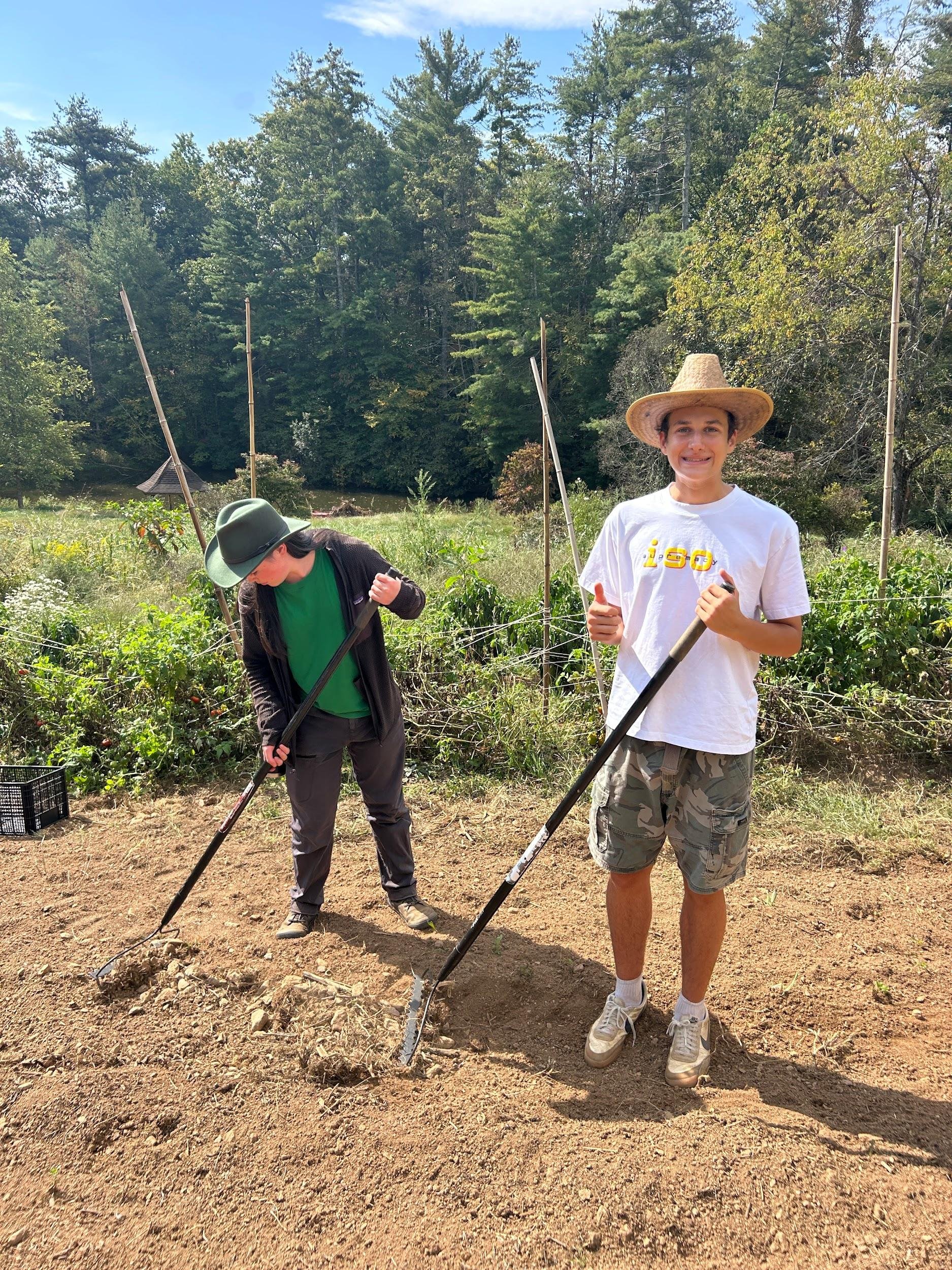 Two young people planting seedlings in a garden with a background of trees and a pond. One is wearing a wide-brimmed hat, a white t-shirt, and camouflage shorts, smiling and holding a gardening tool. The other is wearing a green hat, black jacket, an