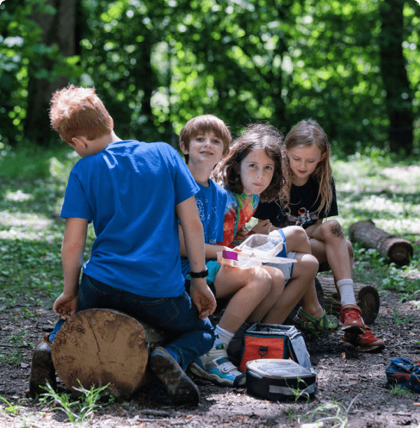 Four children sitting on logs in a forest, with backpacks and snacks.