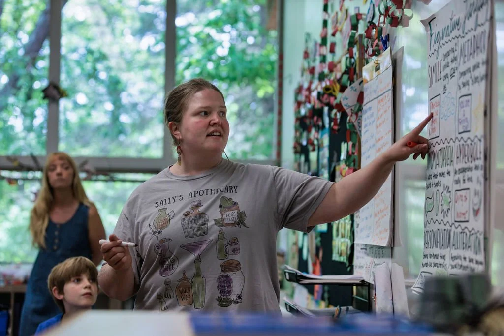 A girl with braided hair pointing at a large colorful poster with handwritten notes in a classroom. Two other children, a boy and a girl, are watching her. The classroom has large windows with a view of trees outside.