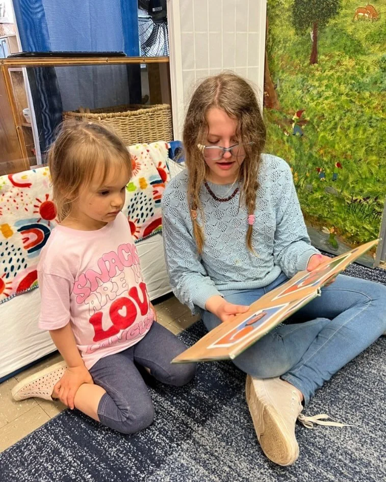a middle schooler with hair in braids reads a picture book to a preschool buddy