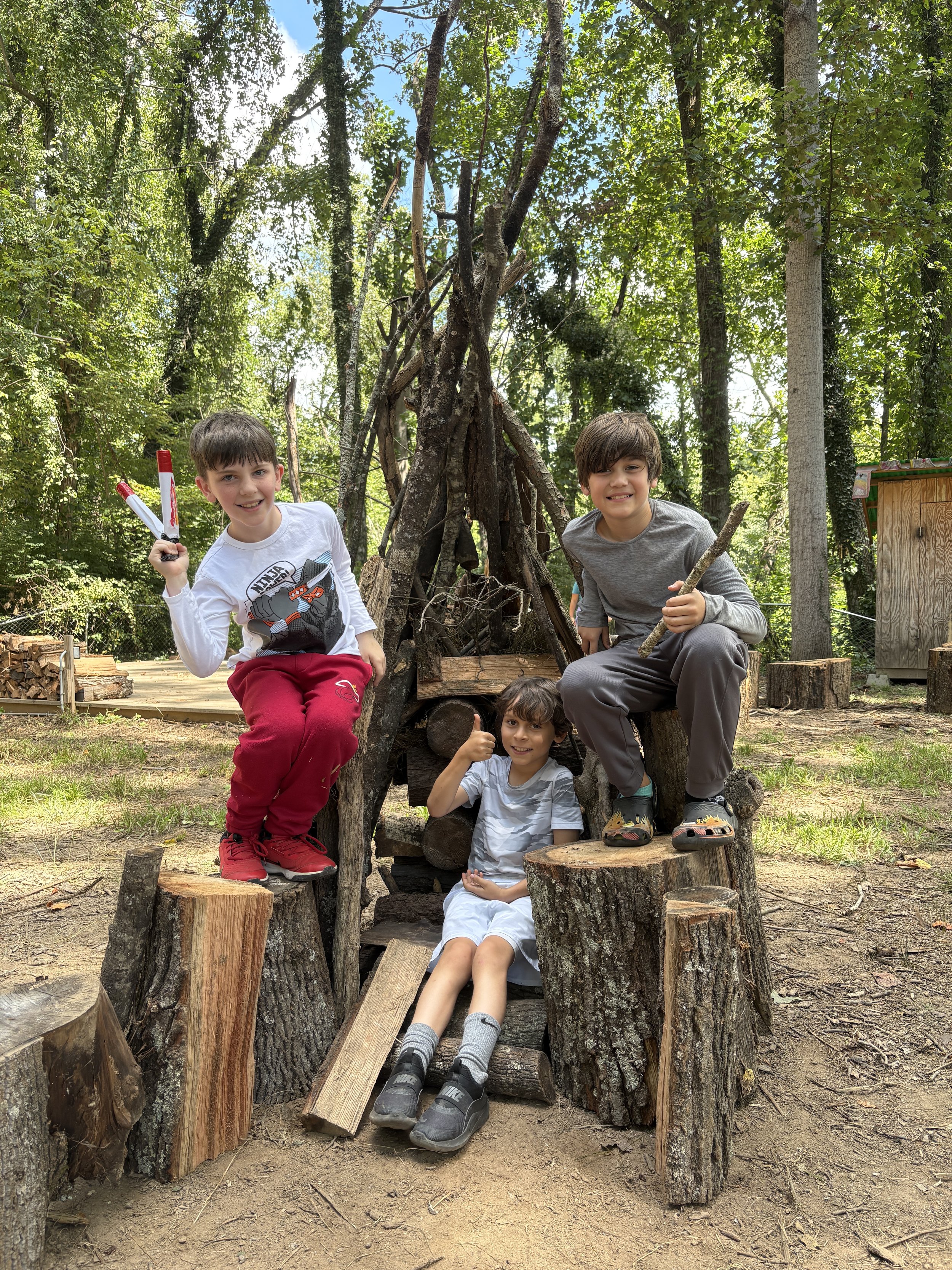 Three boys playing outdoors in a wooded area with a makeshift shelter made of logs and sticks. One boy is sitting on a log, giving a thumbs-up, while the other two are standing or sitting on logs nearby. All appear happy and engaged in outdoor play.