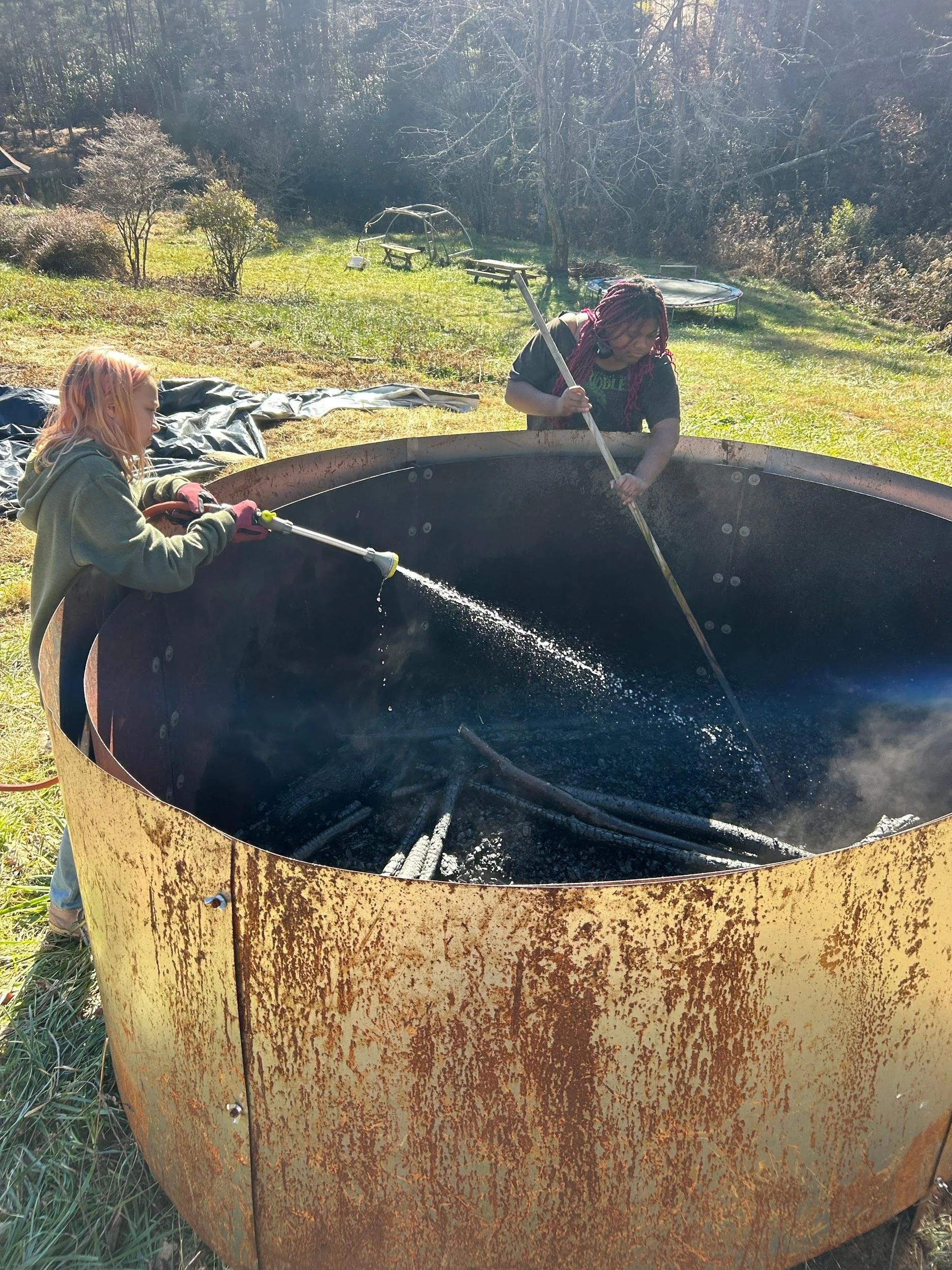 Two people are cleaning a large, circular, rusted metal fire pit outdoors on a grassy area with trees and shrubs in the background.