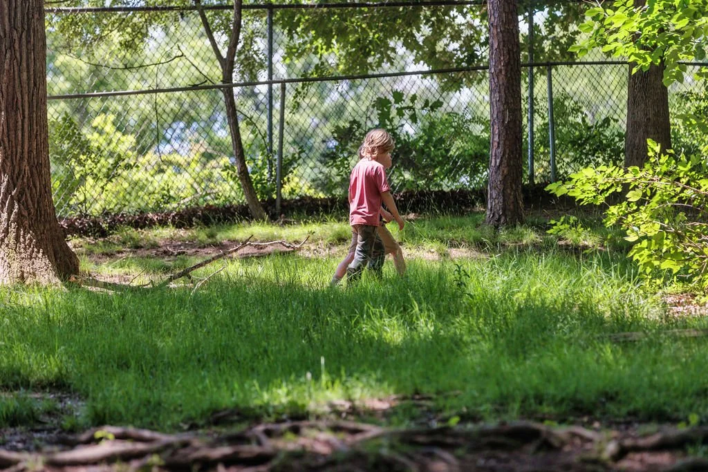 A young girl walking in a grassy outdoor area near trees and a chain-link fence.