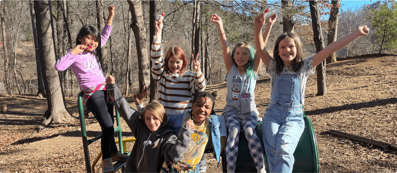 Six girls playing outdoors on a playground, smiling, raising their arms, and having fun in a wooded area.