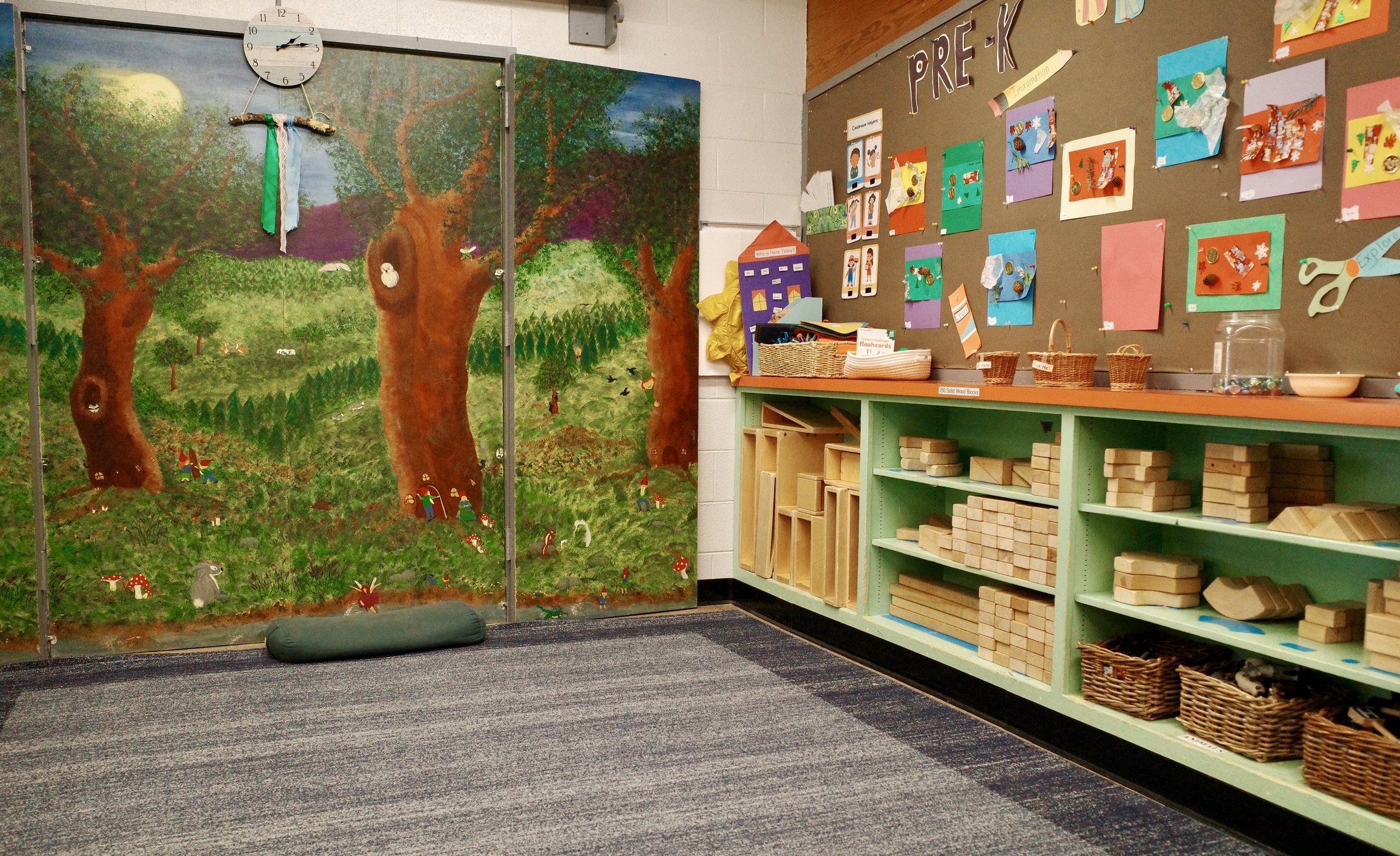 A colorful classroom corner with a painted forest scene on panels, a wall clock, shelves with wooden blocks, and a bulletin board labeled 'PRE-K' displaying children's artwork.