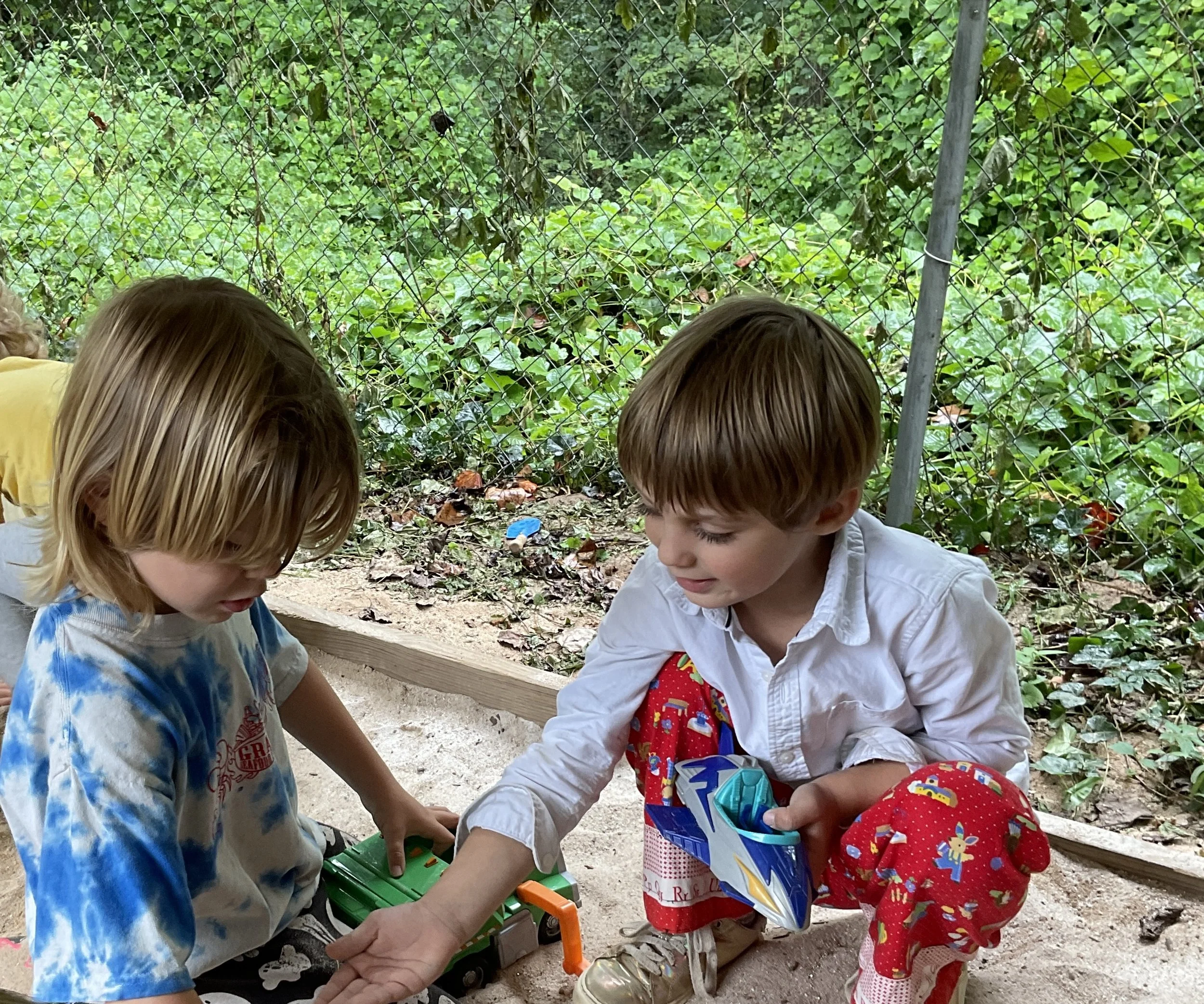 Two young boys playing in a sandbox outdoors, with green foliage and a chain-link fence in the background.