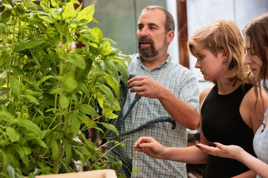 Three people, a man and two high school students, are looking at a large plant with green leaves in a greenhouse or indoor garden.