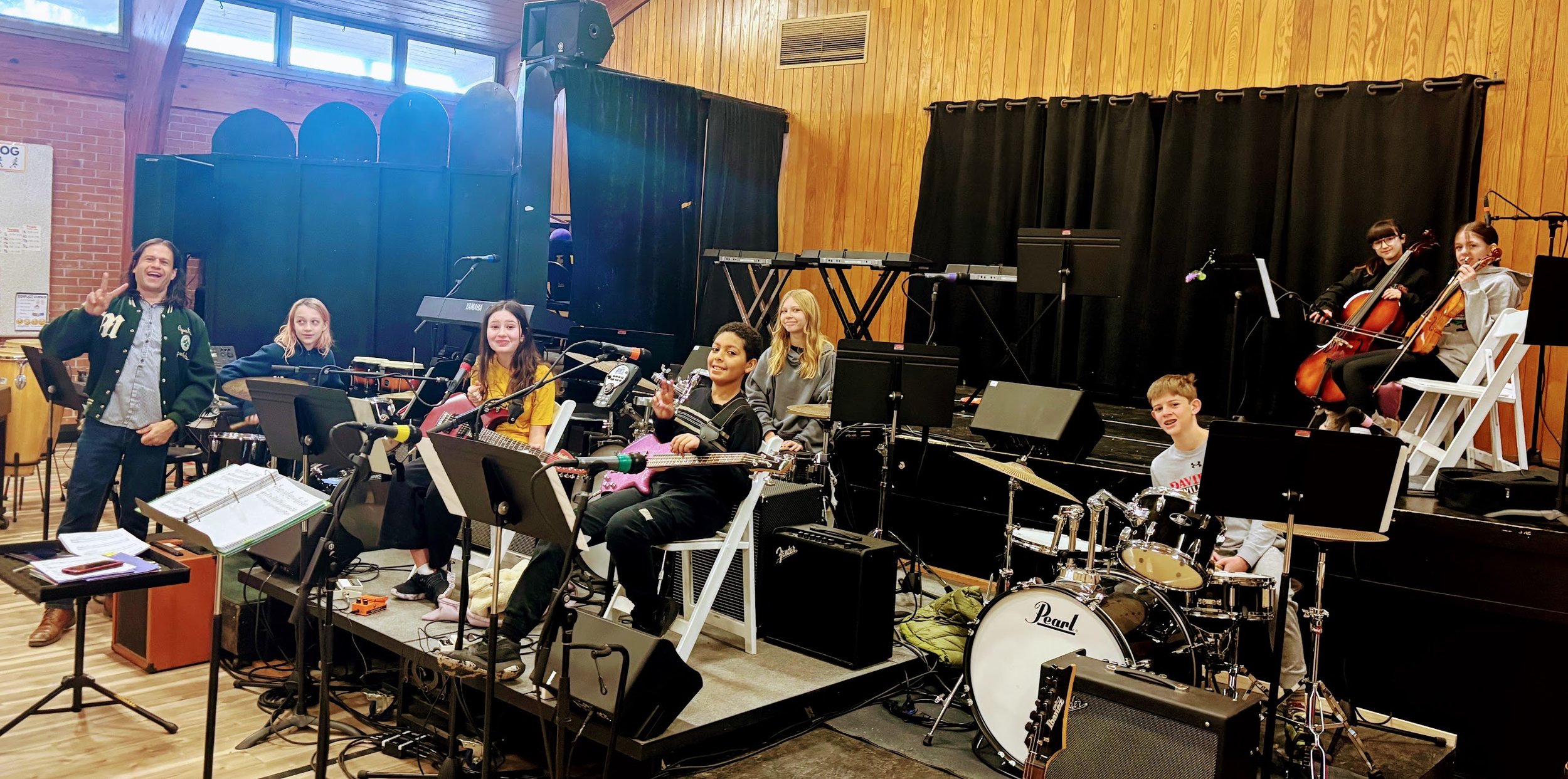 A group of children and a teacher in a school music room, with students holding musical instruments and smiling after a rehearsal or performance.