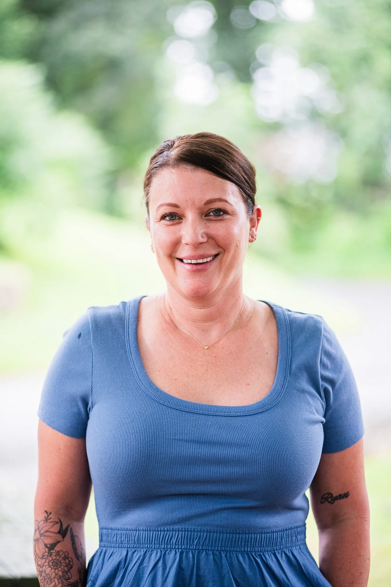 Smiling woman with short dark hair, wearing a blue short-sleeve dress, standing outdoors with blurred greenery background.