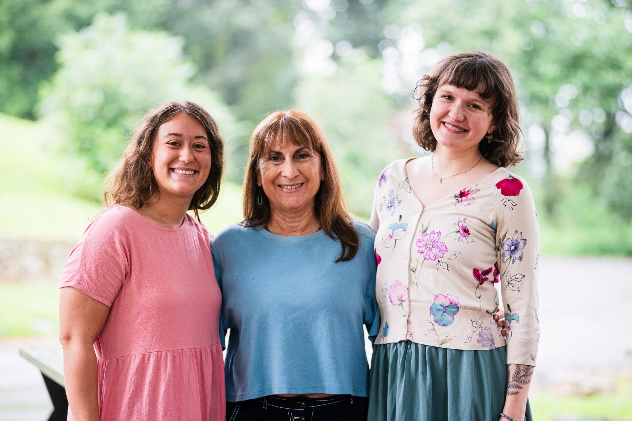 Three women standing outdoors, smiling. From left to right: a woman in a pink dress, a woman in a blue top, and a woman in a floral blouse and teal skirt. Green trees and a blurred background
