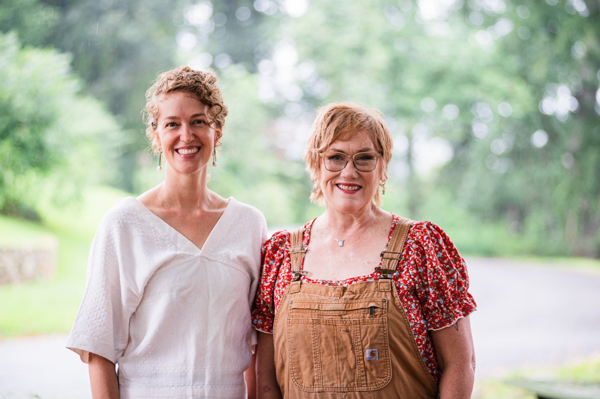 Two women standing outdoors with greenery in the background, smiling at the camera. The woman on the left has curly hair and is wearing a white top, the woman on the right has short hair, glasses, and is wearing a red floral blouse with a brown overa