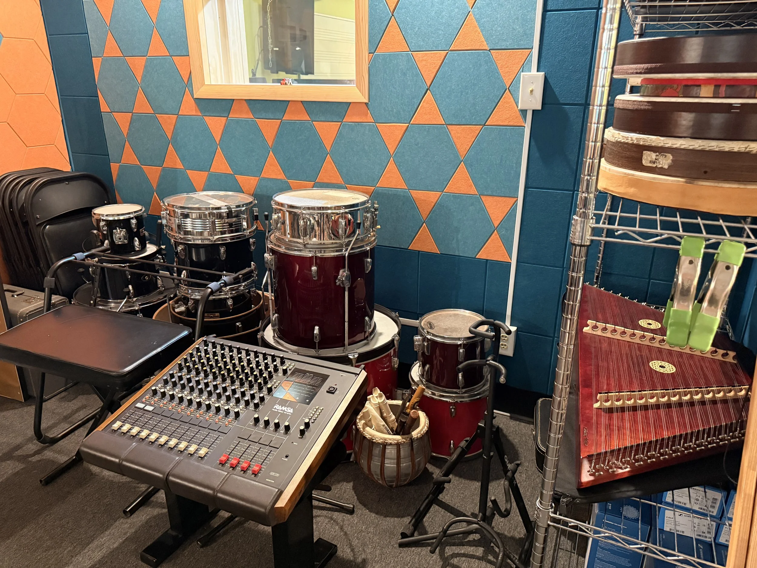 A room with percussion instruments, a sound mixer, and stacked tray-like instruments on a metal shelf, set against a colorful tiled wall.