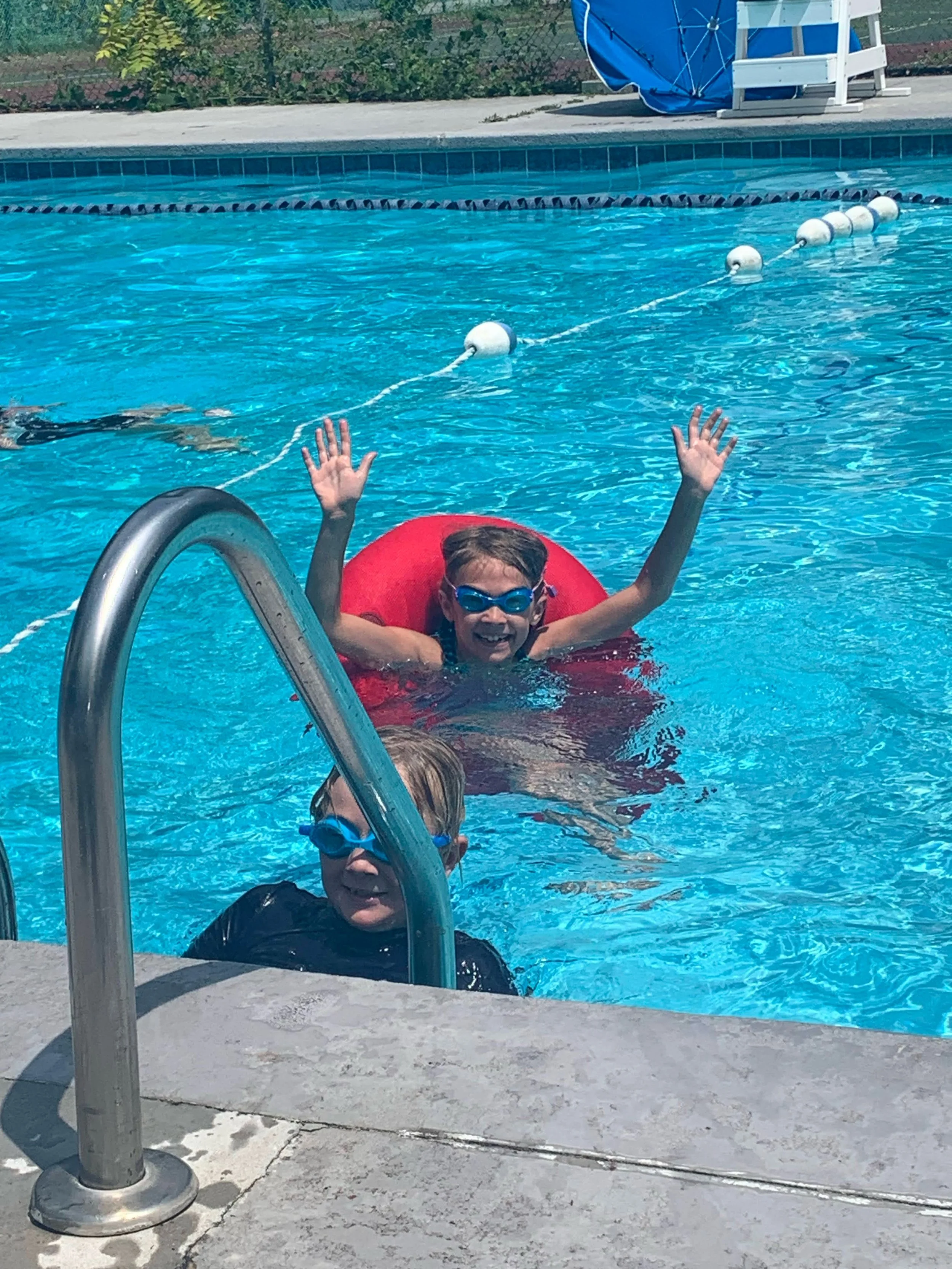 Two children in a swimming pool, one with goggles near the ladder, and another smiling with arms raised, wearing goggles and floating on a red float.