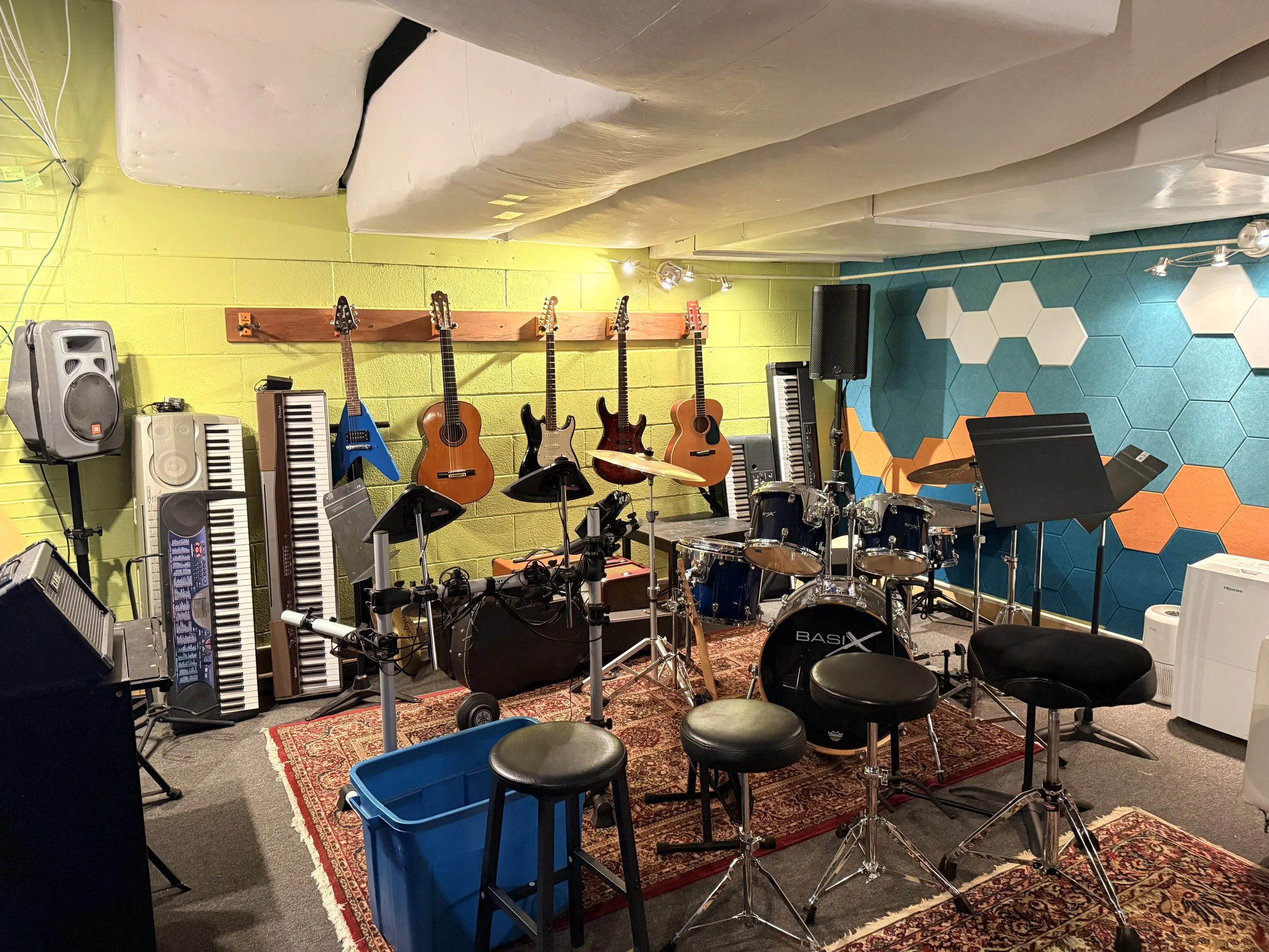 Music practice room with various instruments including guitars hanging on a yellow wall, a drum set, and electronic keyboards. The room has colorful wall panels, a patterned rug, and chairs with music stands.