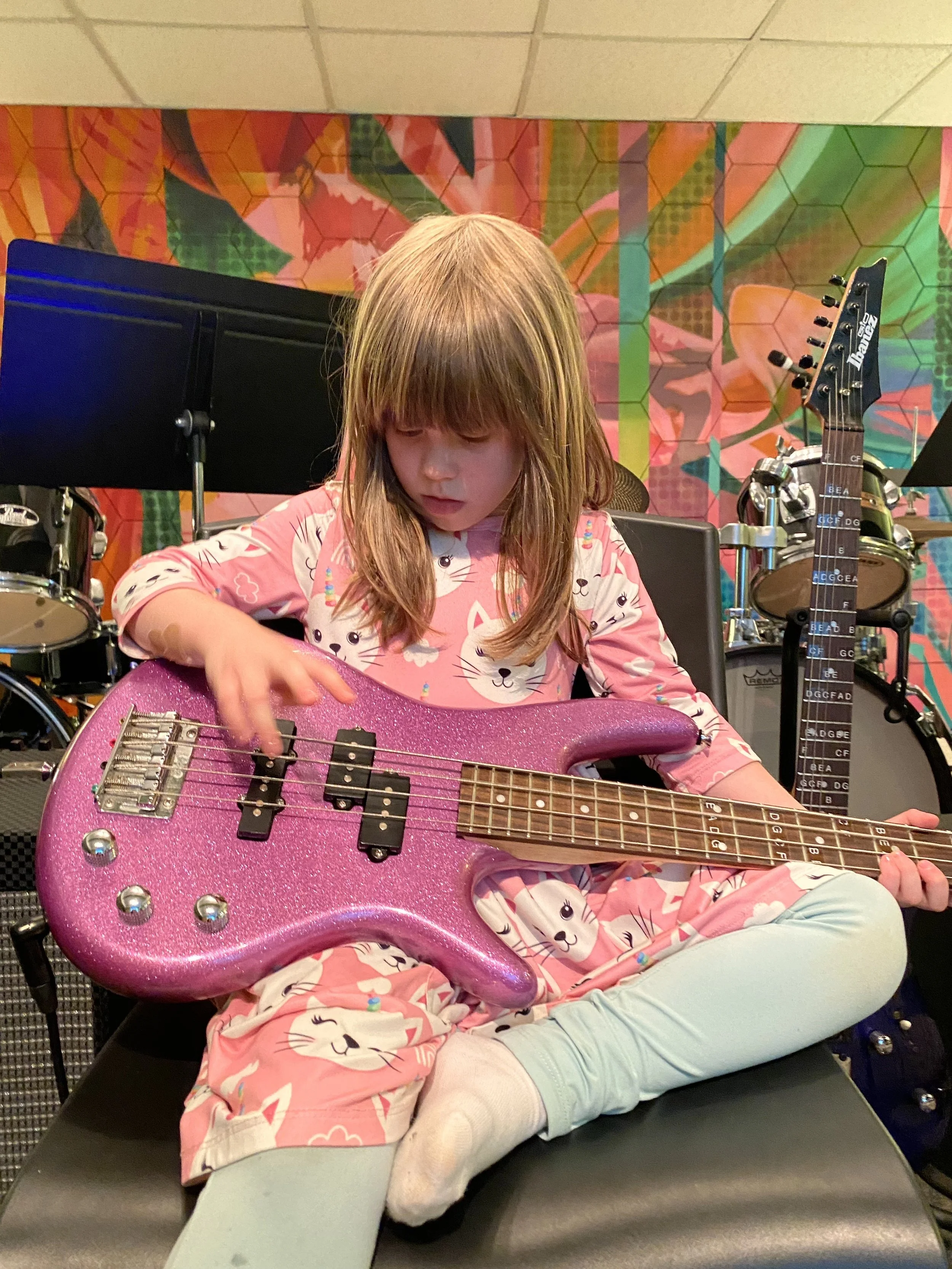 A young girl is sitting cross-legged on a bench playing a pink glittery electric bass guitar in a music room with colorful graffiti-style wall art.