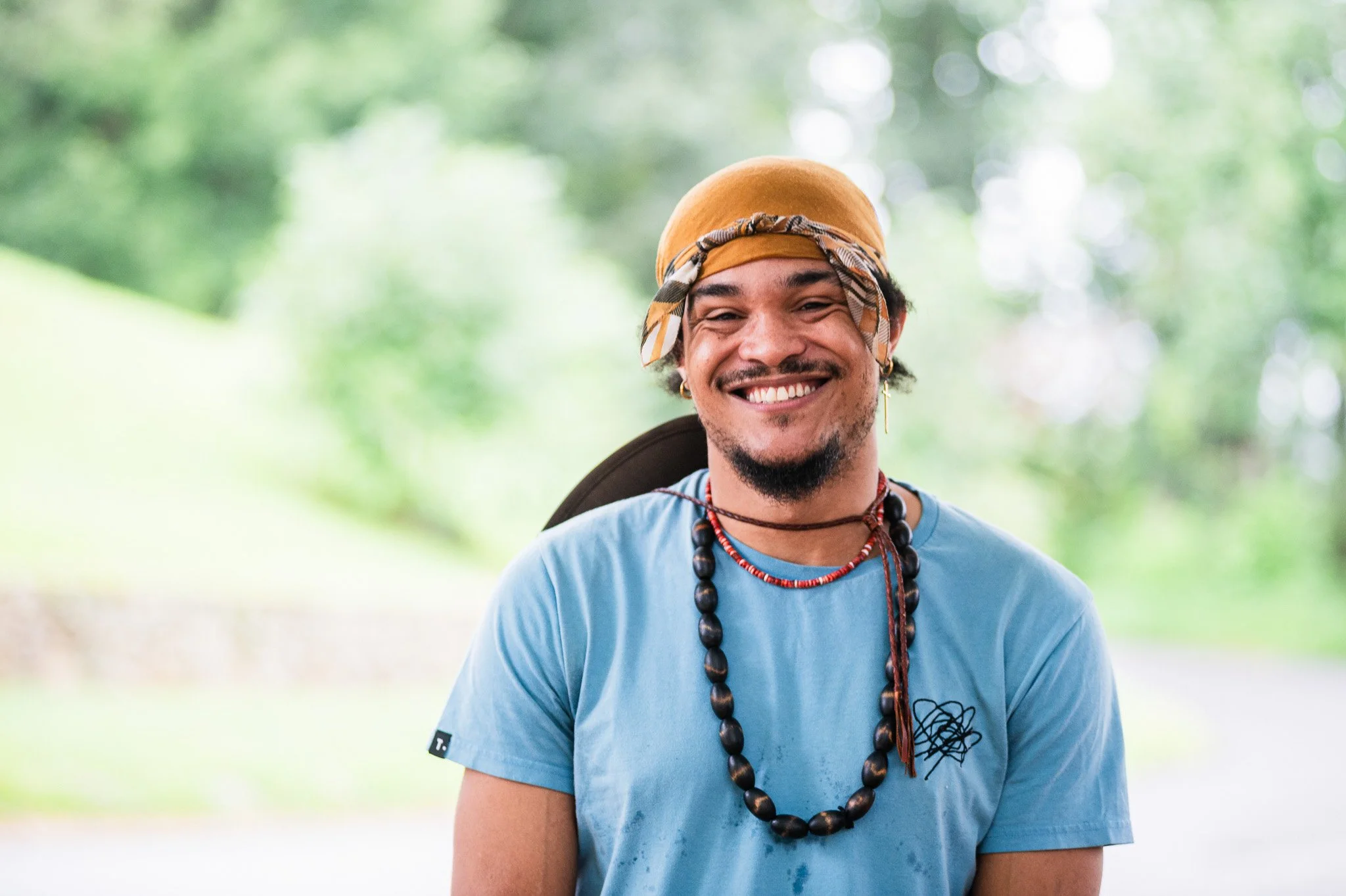 Smiling man wearing a brown bandana and a light blue shirt outdoors with greenery in the background.