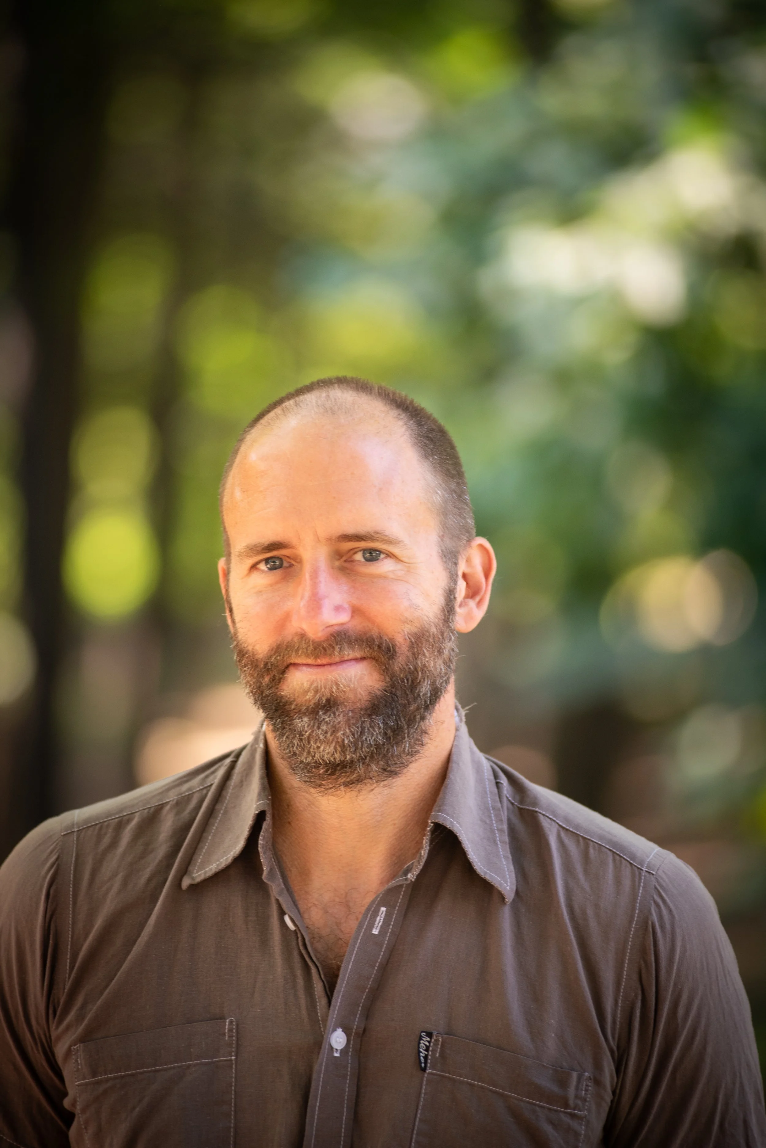 Brown-bearded man in a brown button up shirt standing in front of a forested background