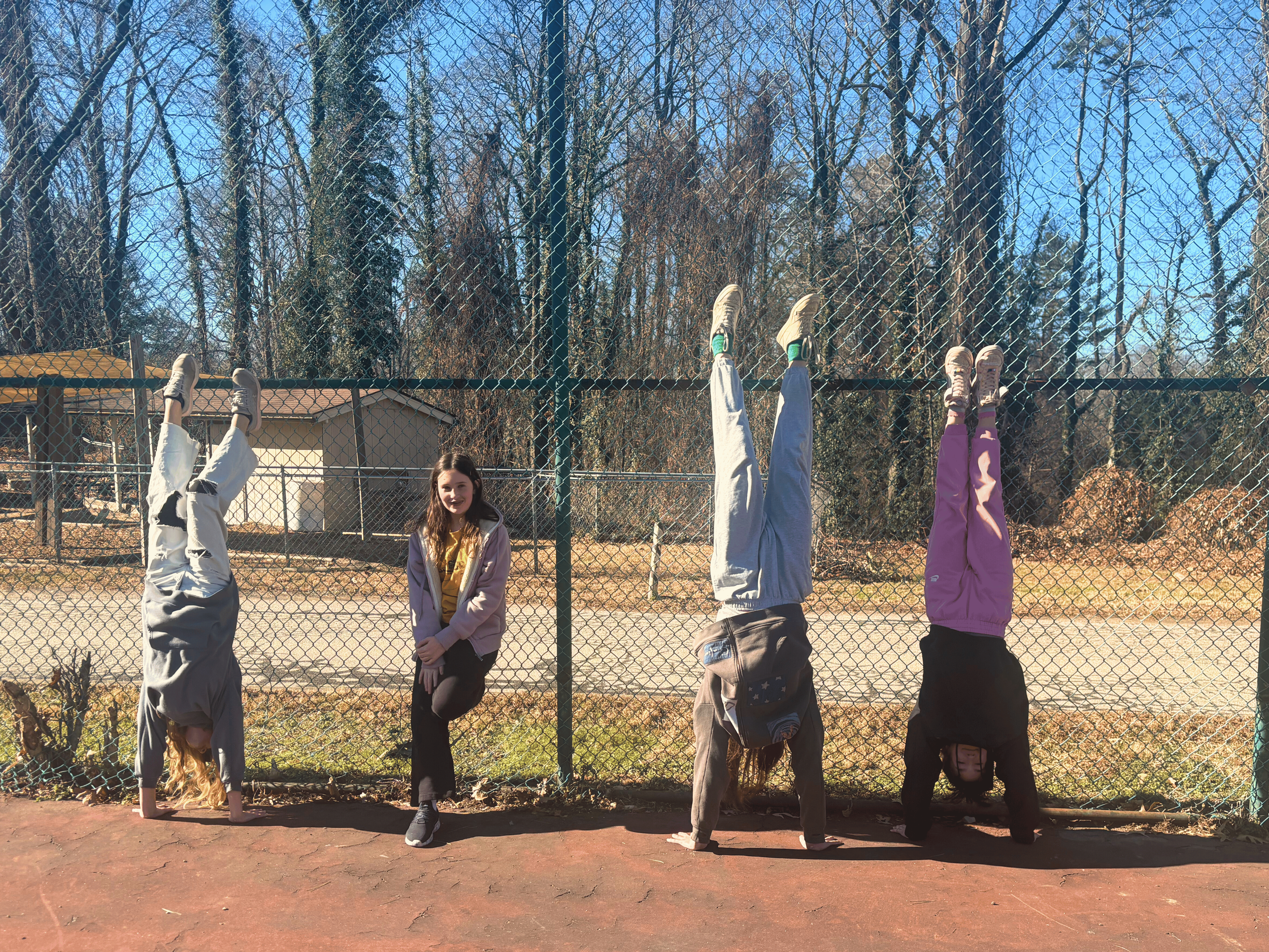 Four young girls practicing handstands against a chain-link fence on a sunny day, with a girl standing in the middle smiling. Behind the fence are trees and a small building.