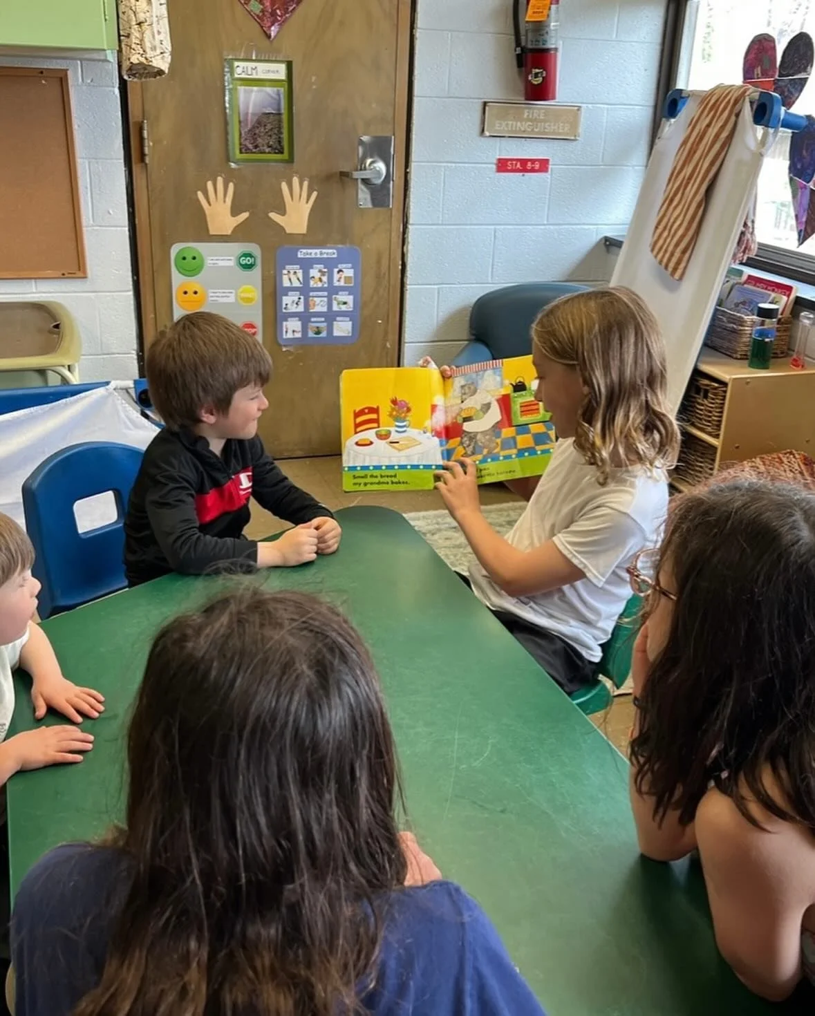 a middle schooler in a white shirt reads bread and jam for Frances to a group of preschoolers at a green table